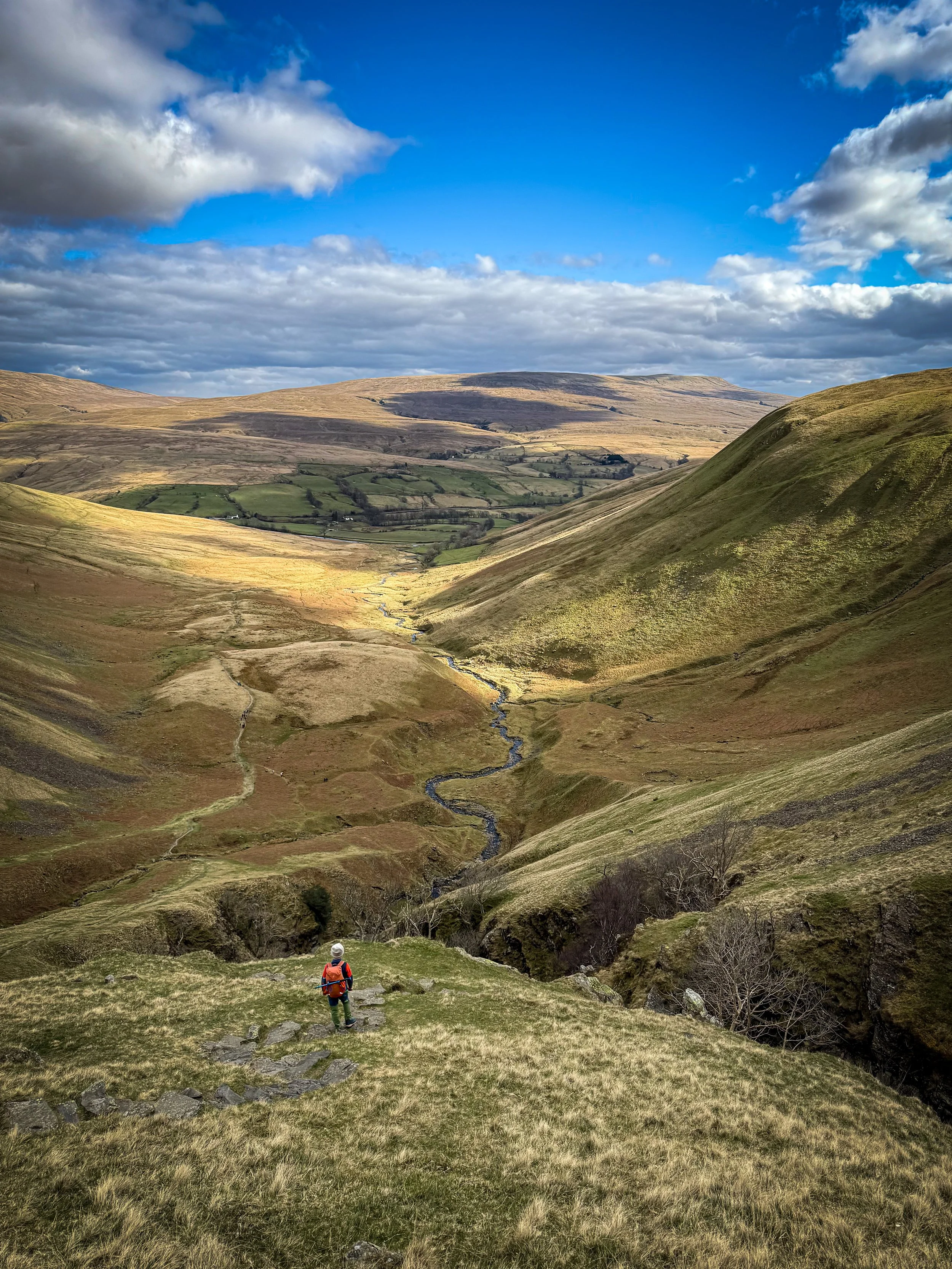 Child standing at the top of a steeply sided gorge with a river snaking in the distance with fells in the background