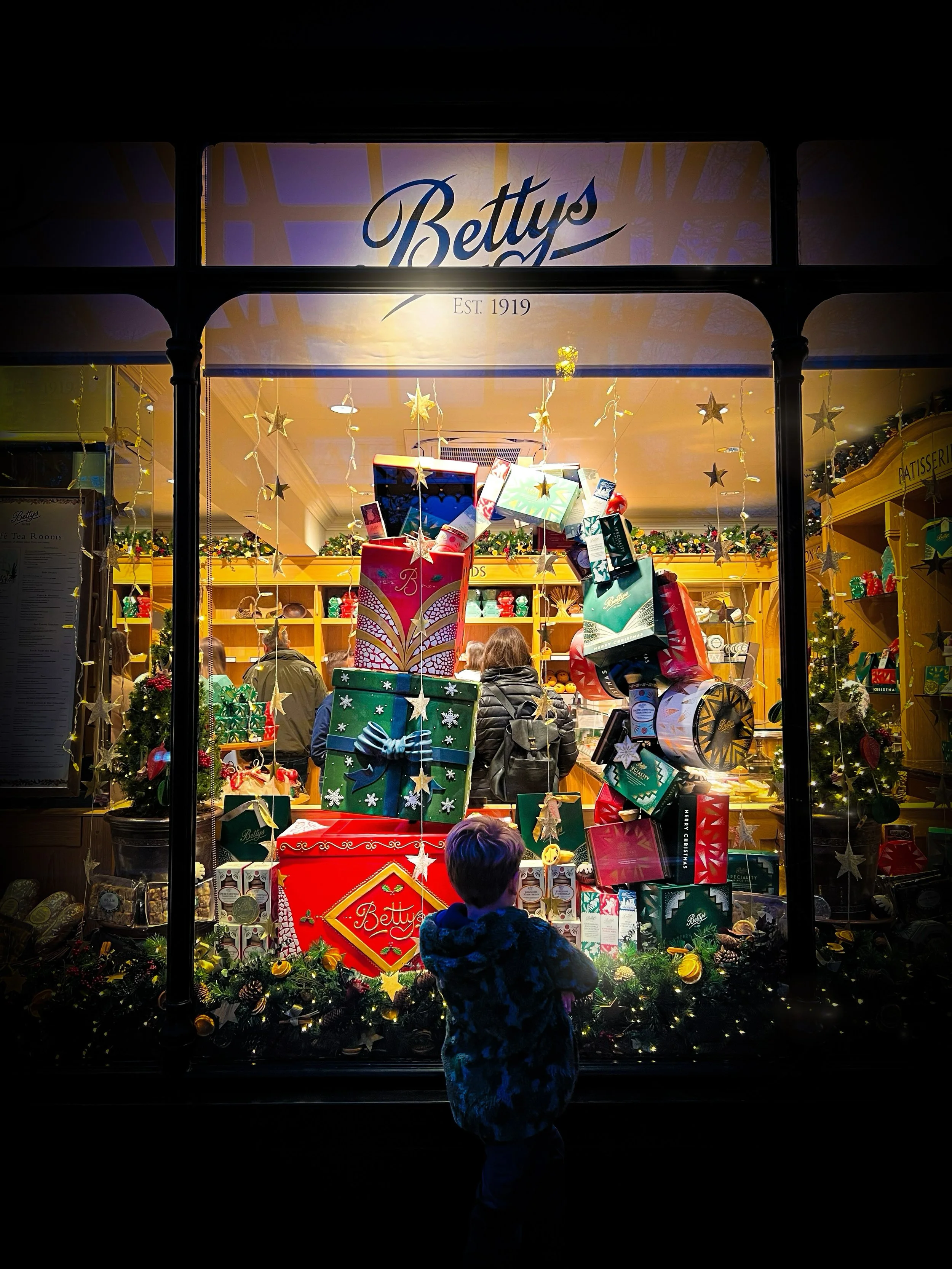 Child standing in front of a shop window thats decorated for christmas with gifts piled up in multi colours and the word Bettys at the top