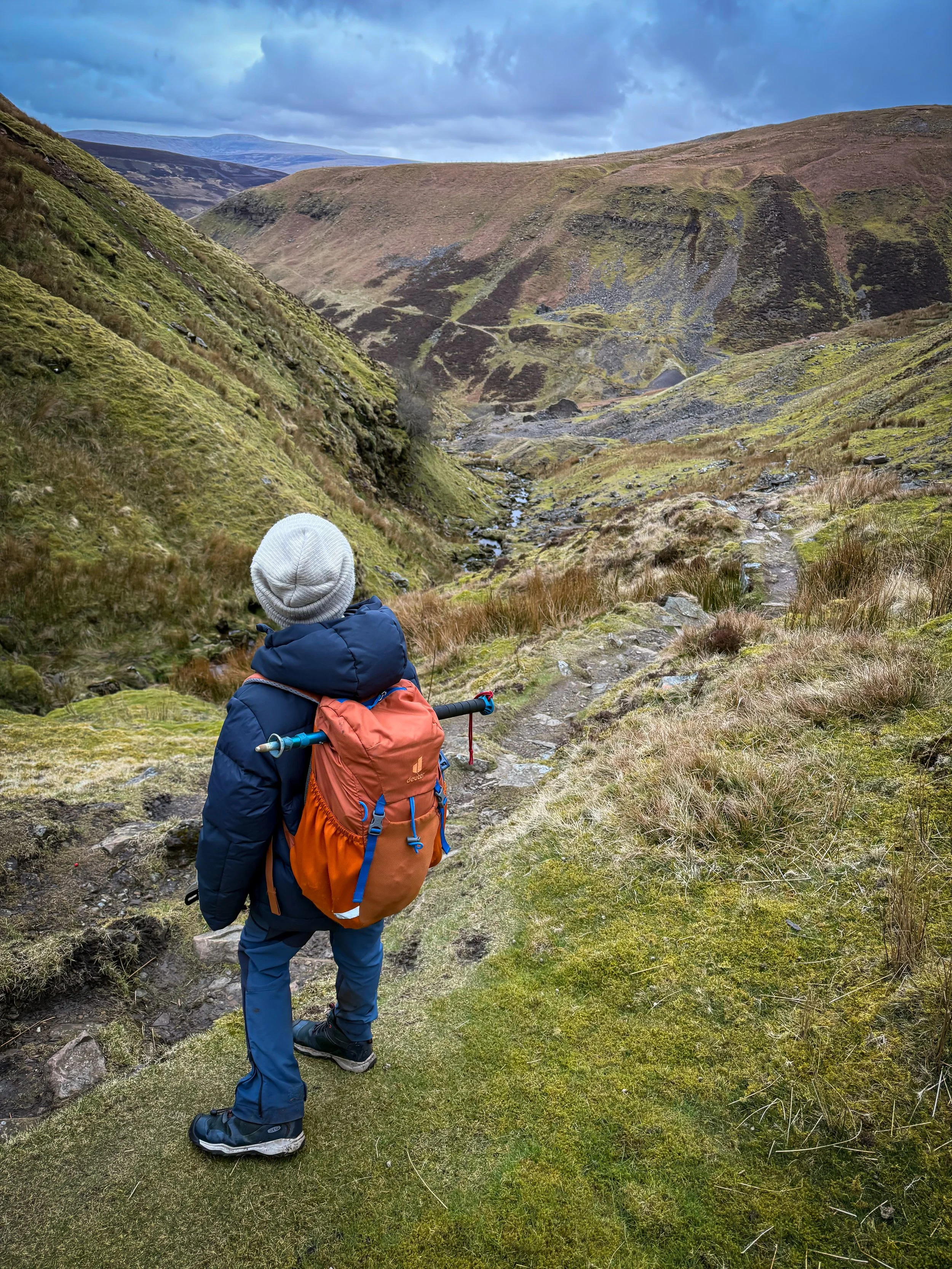 Child standing with orange rucksack and hiking pole looking down a path towards mining ruins