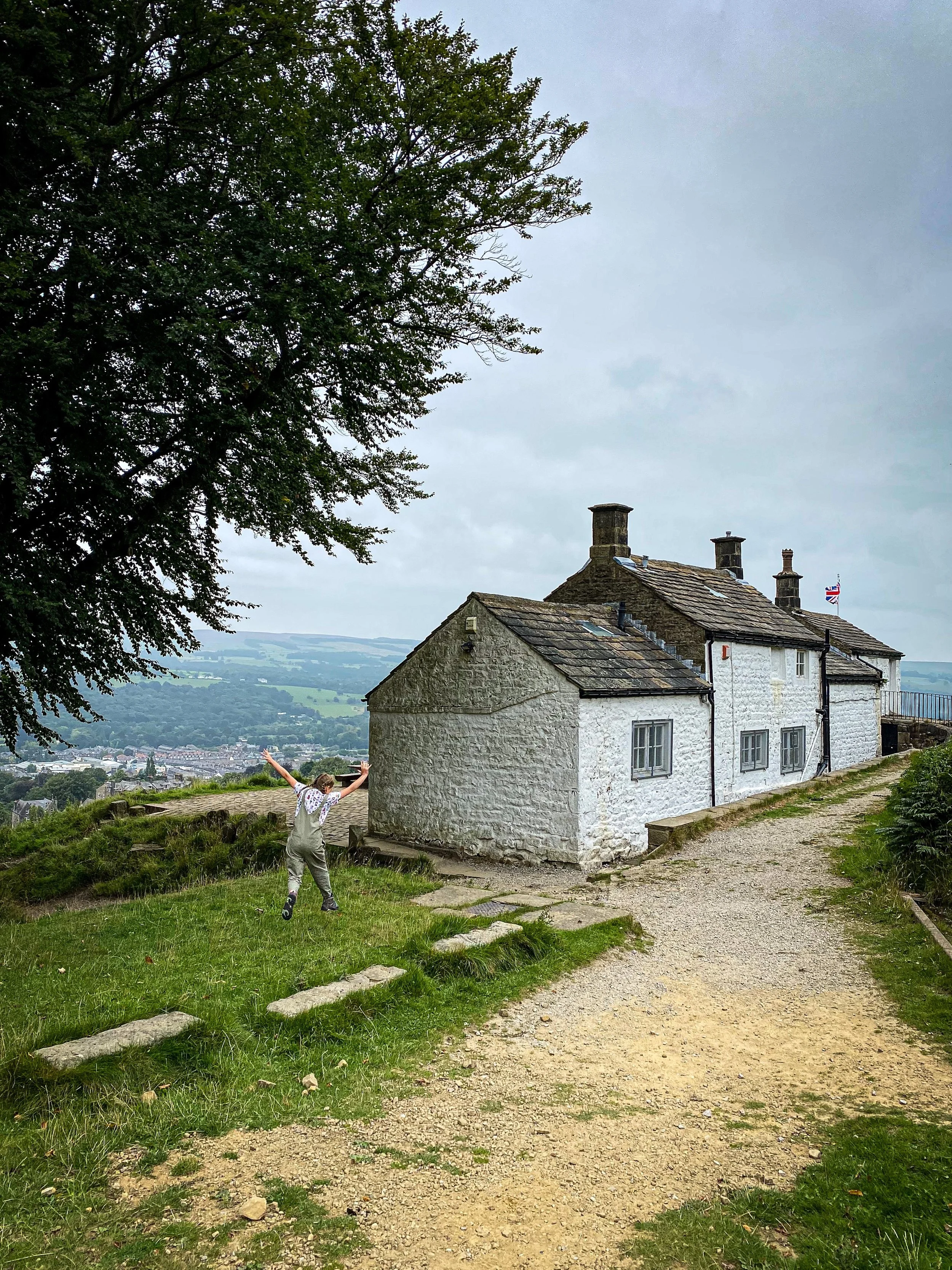 Child jumping in the air next to a white washed house with a tree to the side and views to a town lower down in the valley beyond