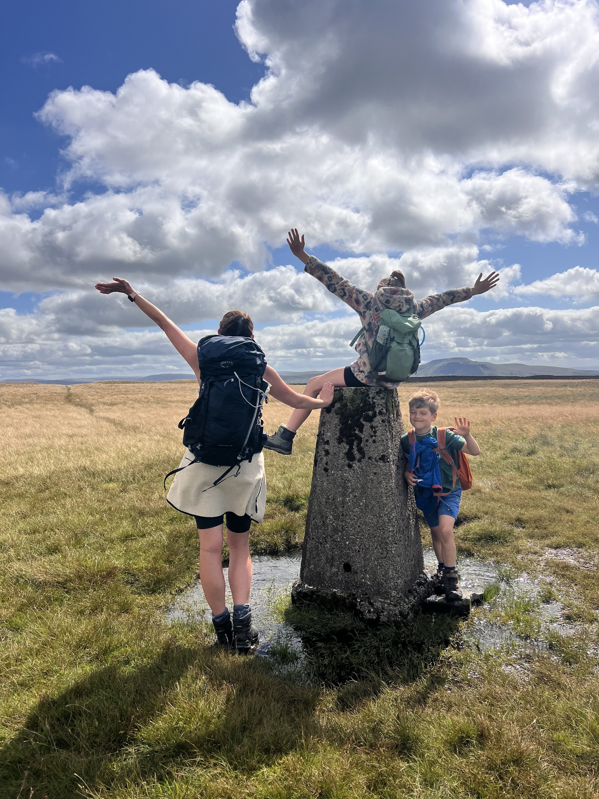 An adult and two children standing at a trig point with arms in the air