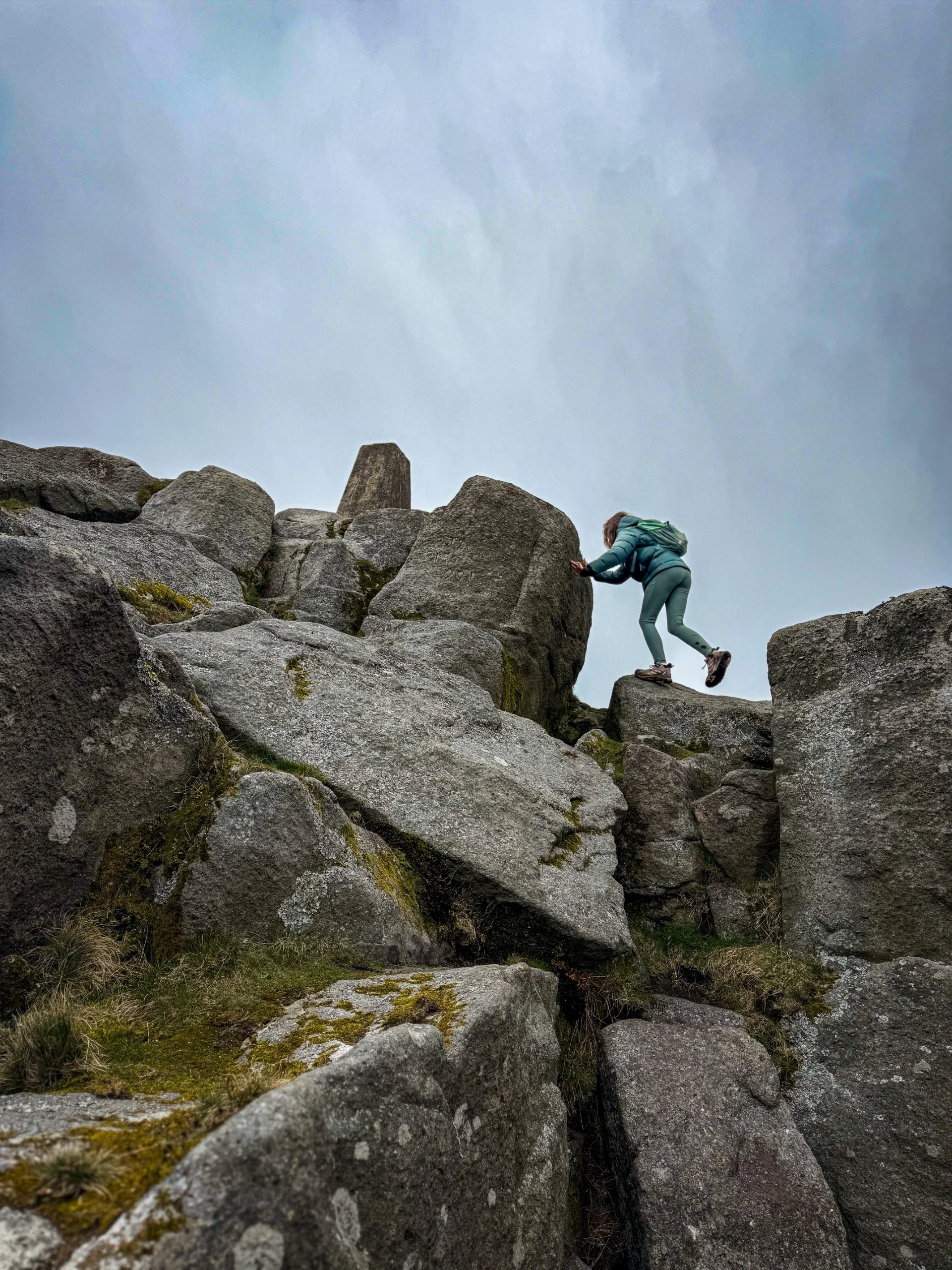 Child scrambling up rocks to reach a trig point under a grey moody sky