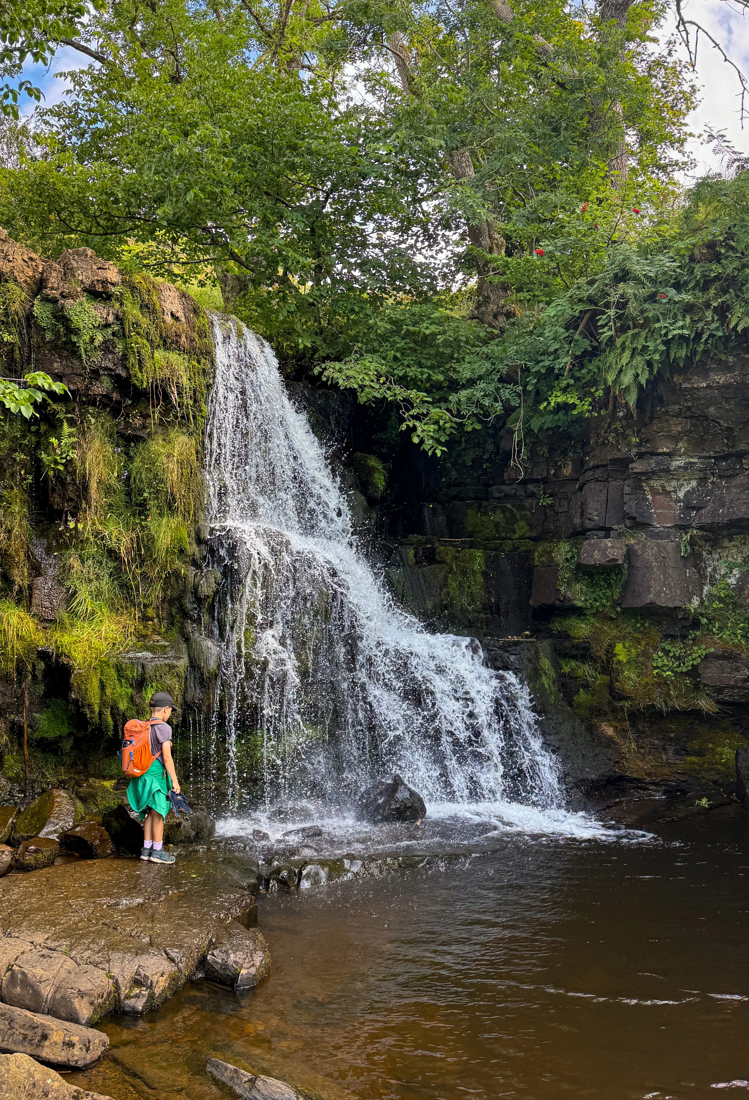 A child standing next to a large waterfall with a backpack on an hiking clothes in summer
