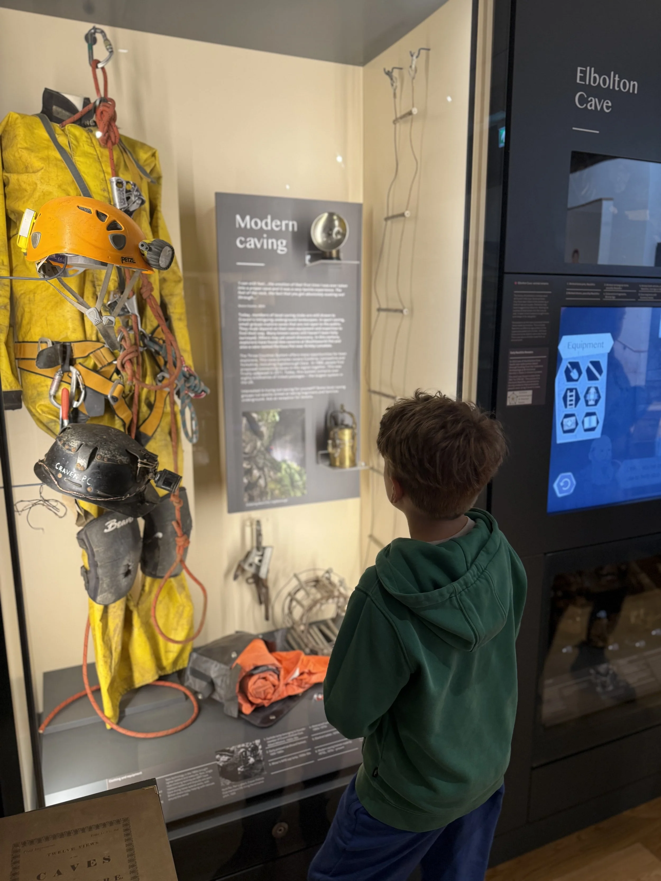 Child looking at a caving exhibit with a caving suit, helmets and information board about caving