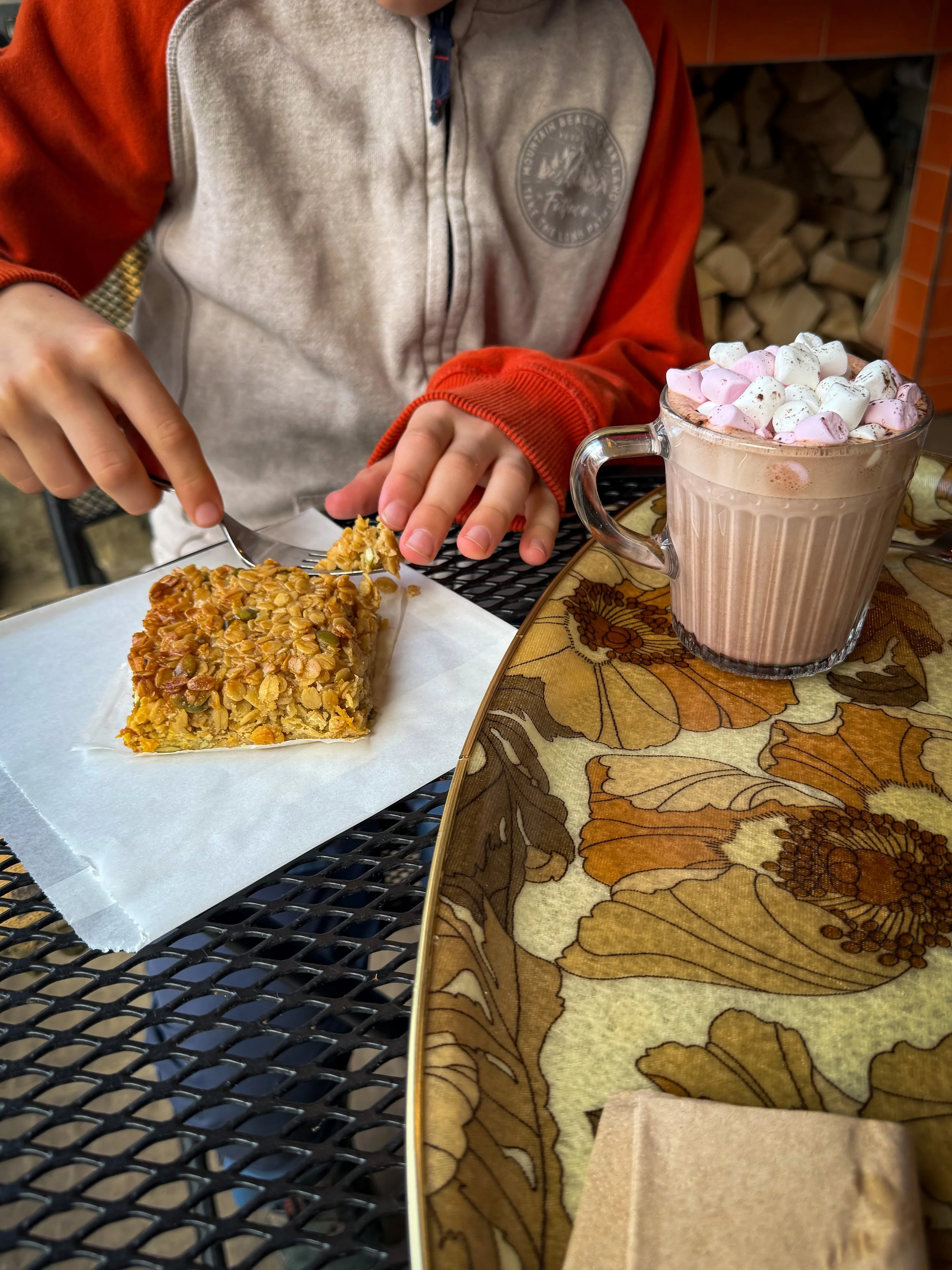 child eating flapjack with a fork which is sat next to a hot chocolate with marshmallows