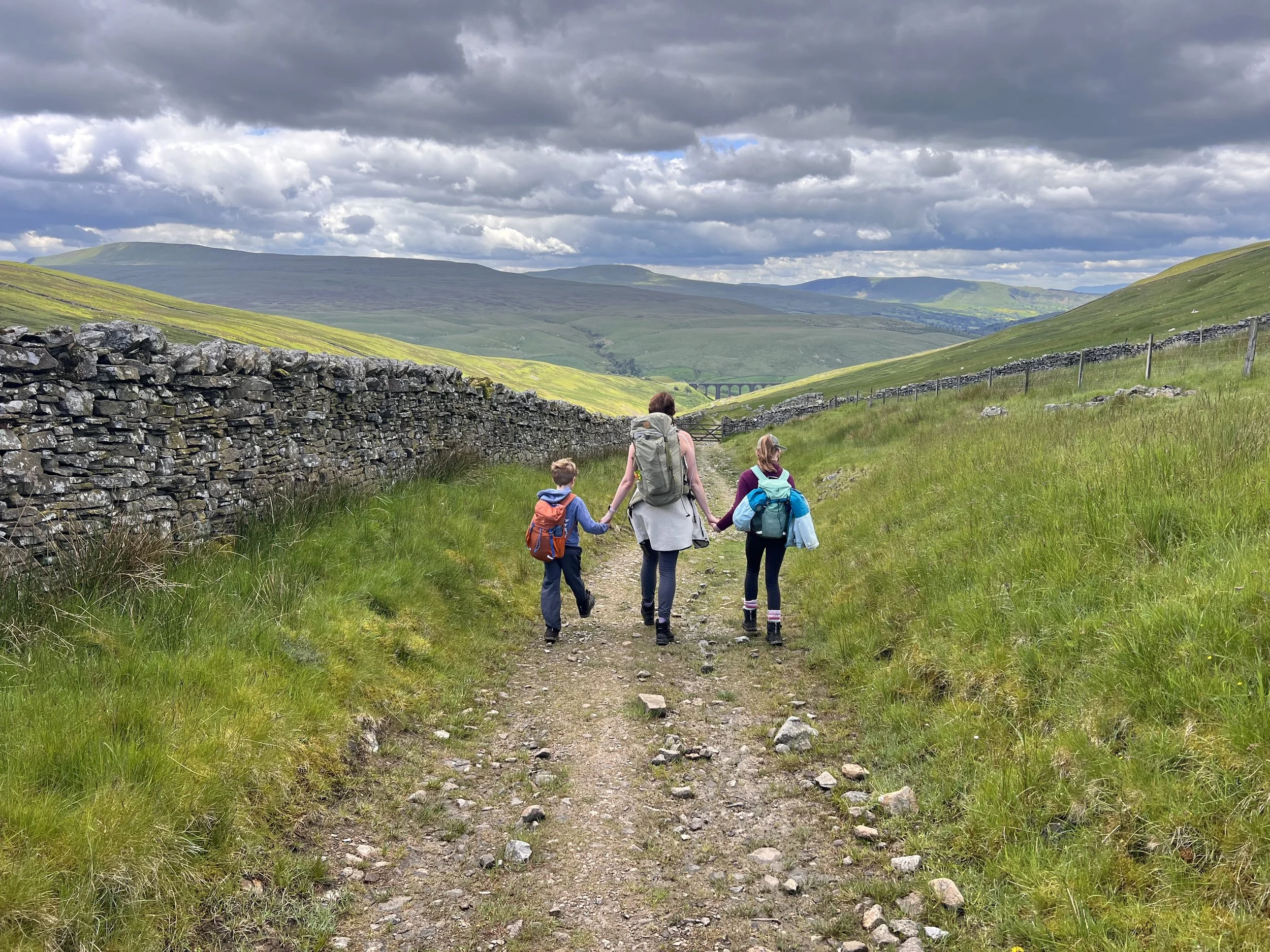 Two children holding hands with their mum each carrying a backpack walking down a stone track with a dry stone wall to the left and a gate and viaduct beyond with mountains ahead