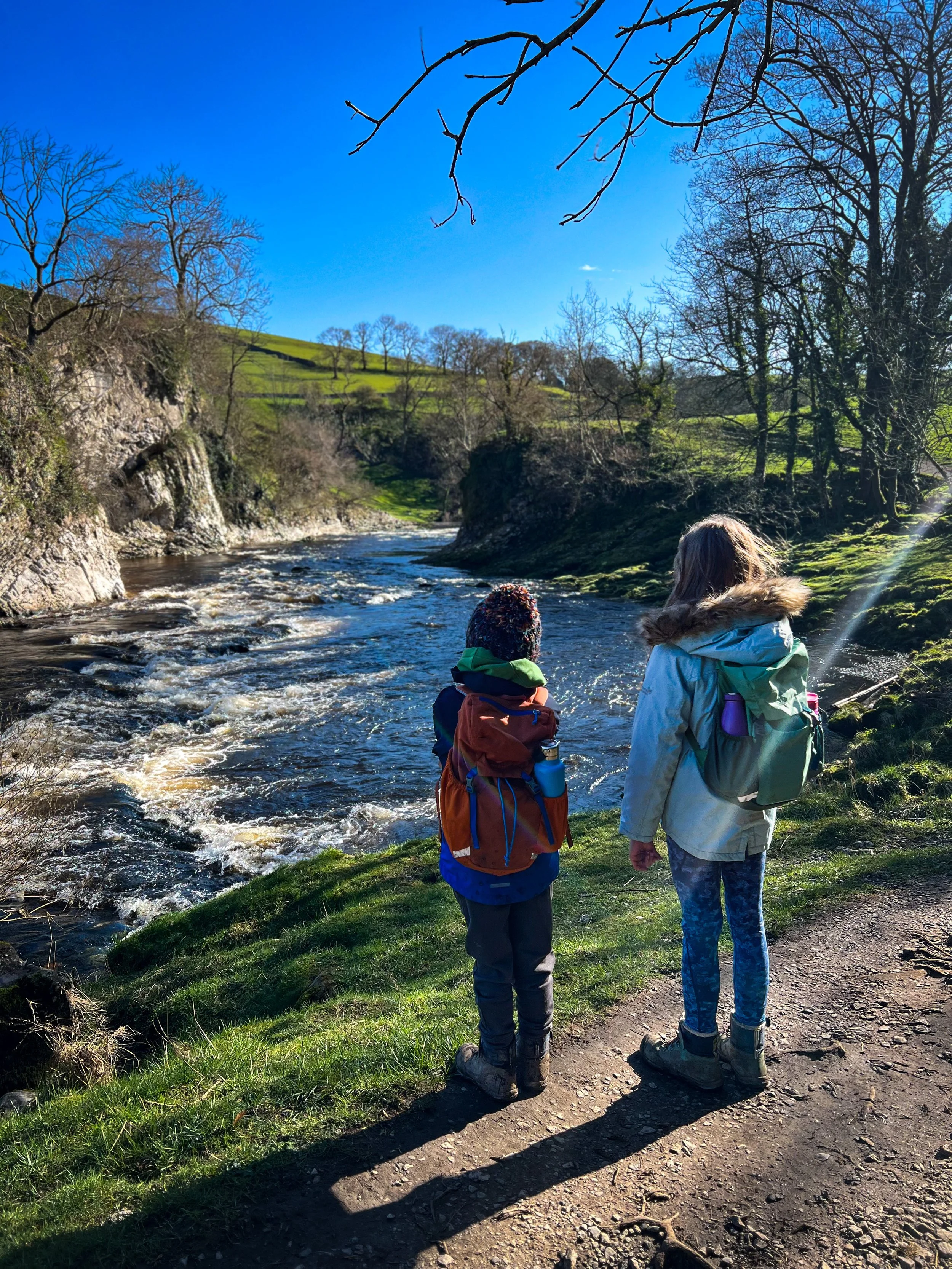 Two children standing looking out towards a shite cliff face and full river in winter under blue sky