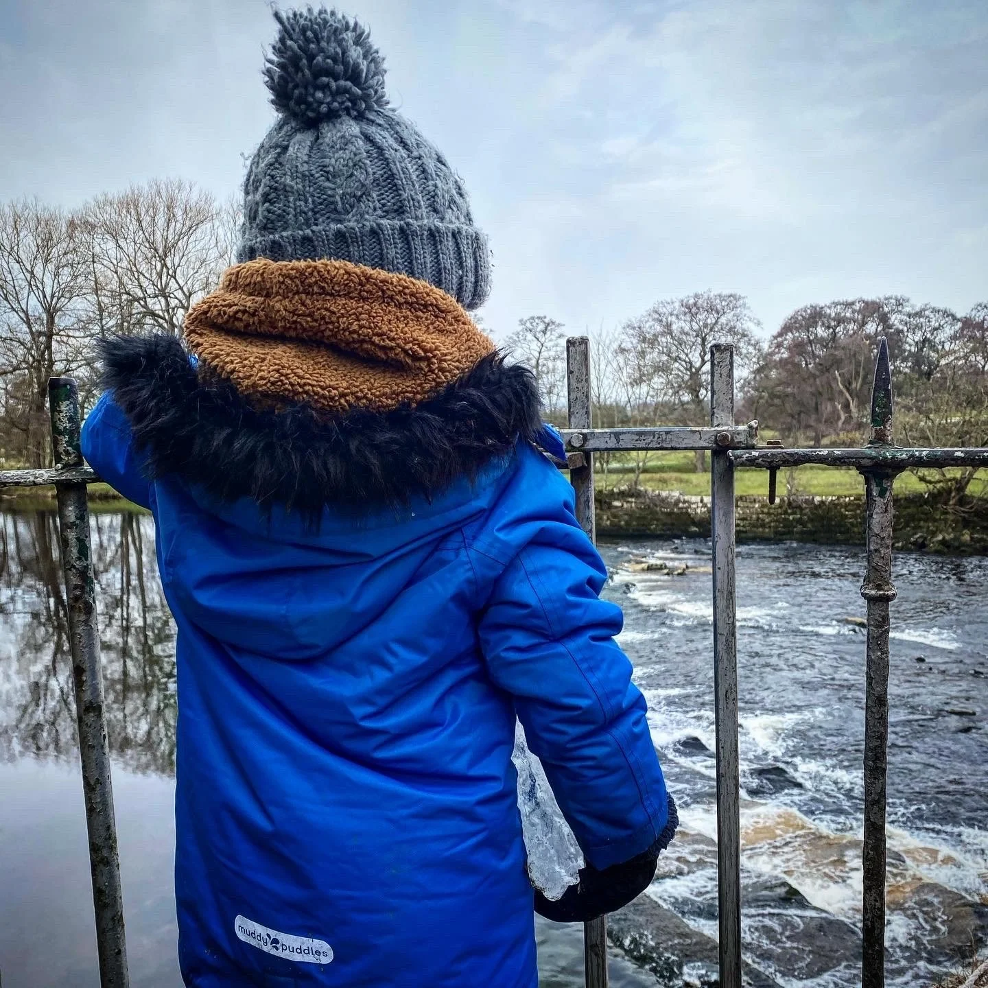 Child standing next to a weir in winter wearing a blue coat and grey bobble hat leaning on iron railings