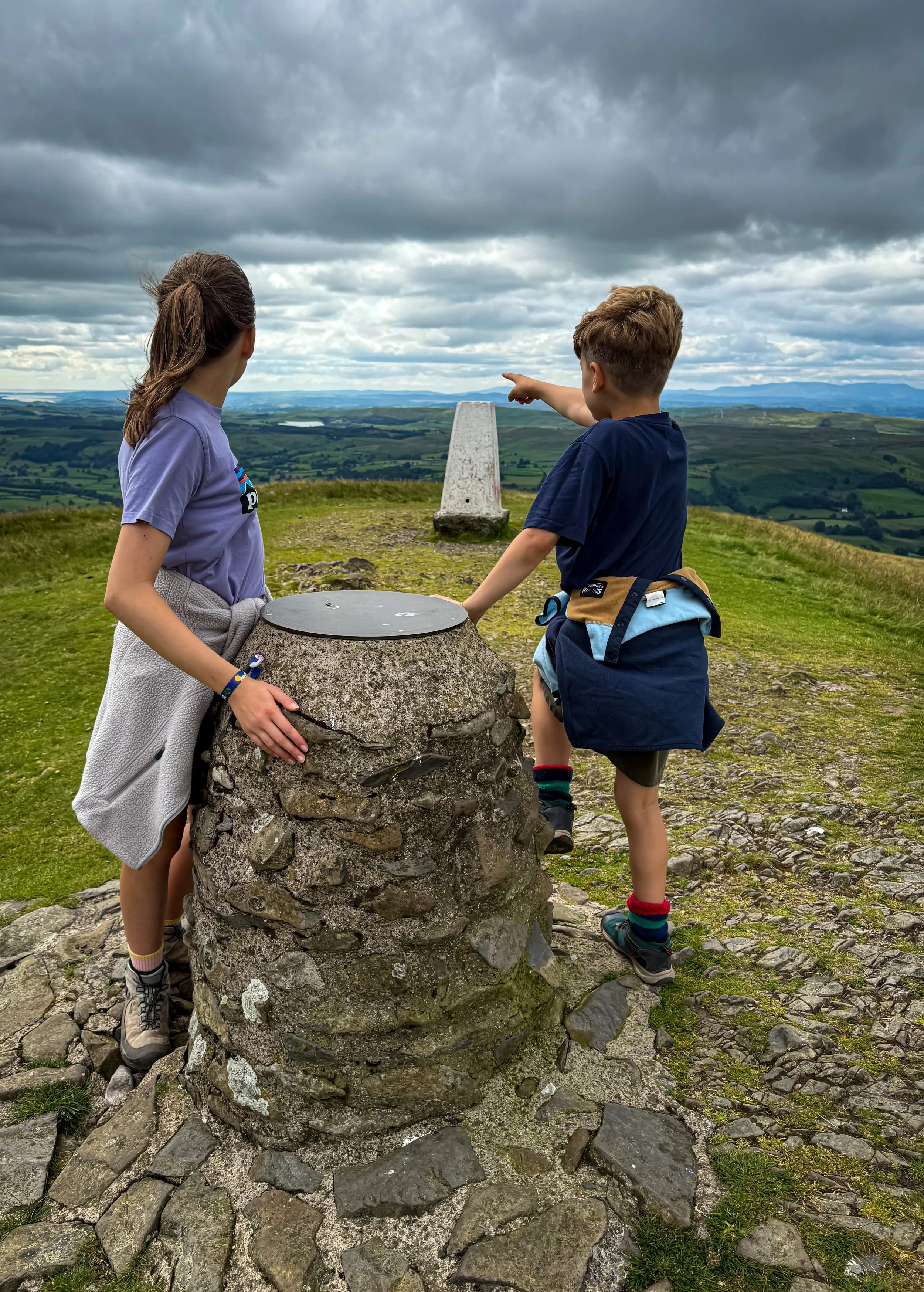 Children on top of a hill with a trig point behind and a view pillar pointing at view