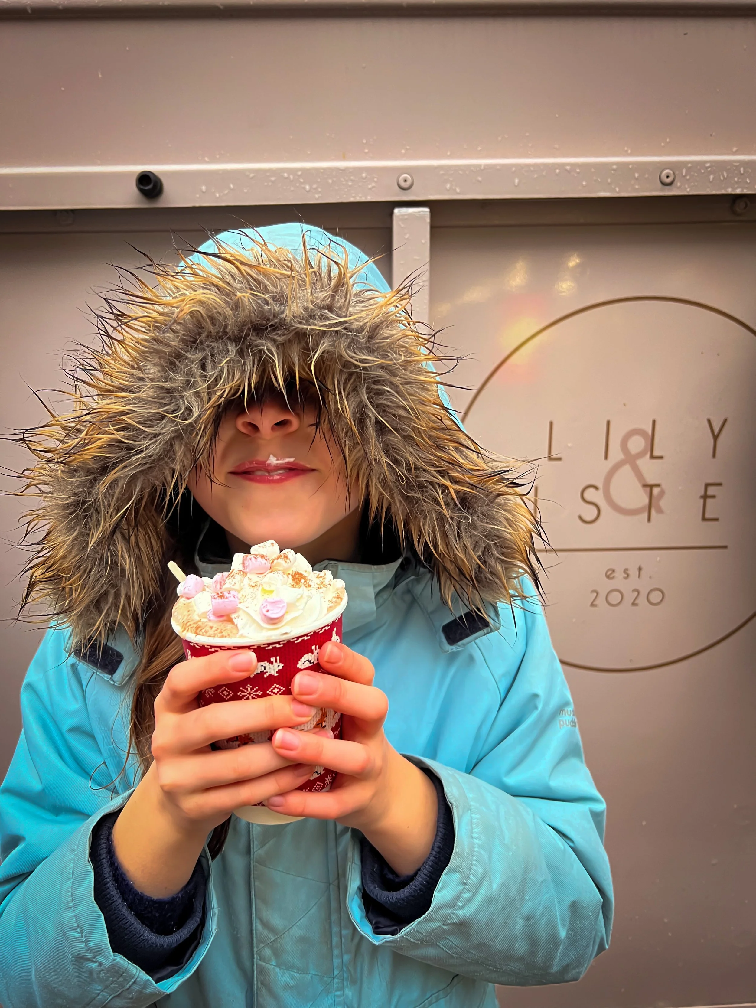 Child wearing a blue parker coat with hood pulled up and over eyes drinking a large hot chocolate with cream and marshmallows smiling in front of a store