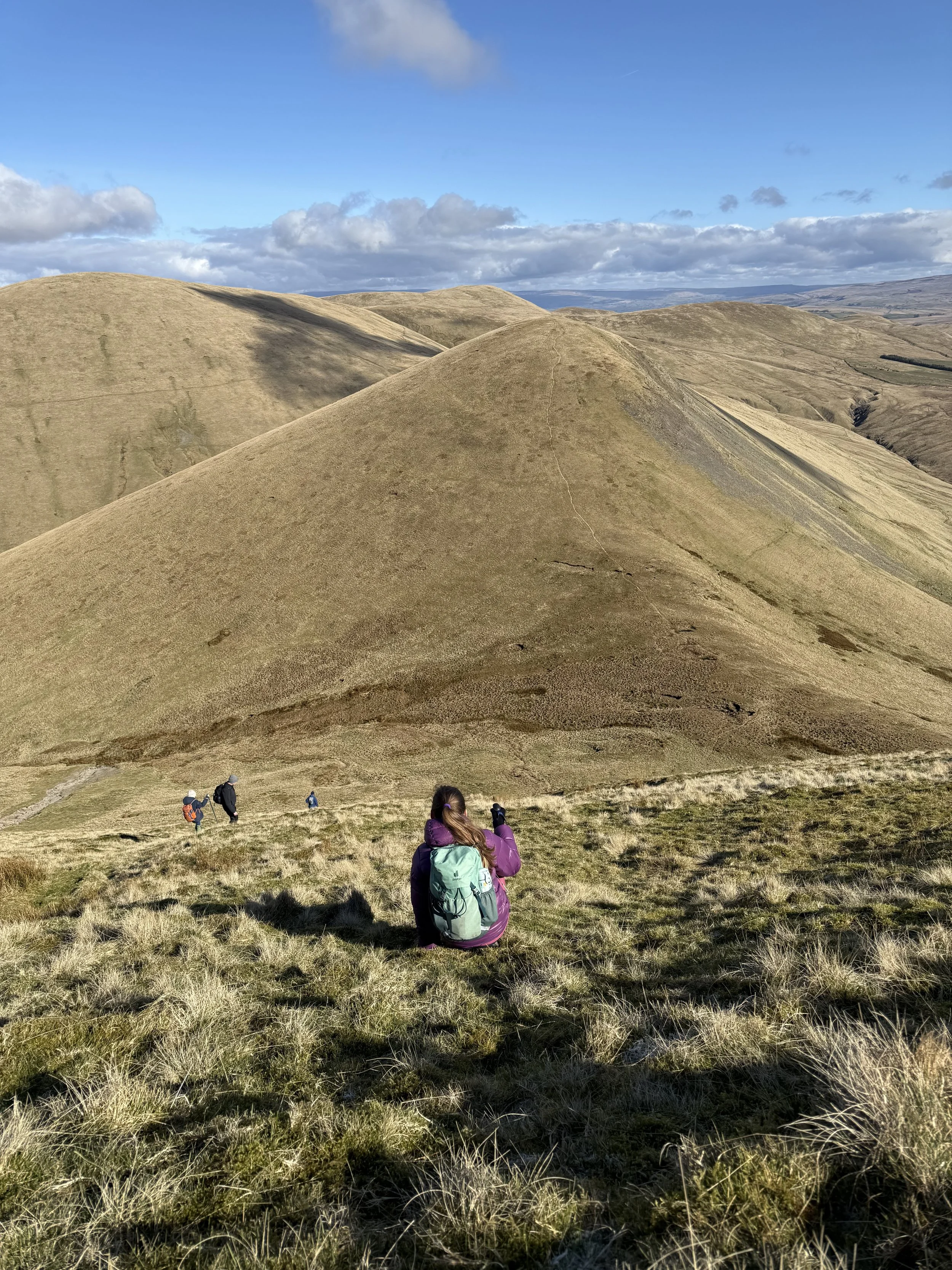 Child sat on the grass of a steep descent from a mountain with grassy mountains beyond