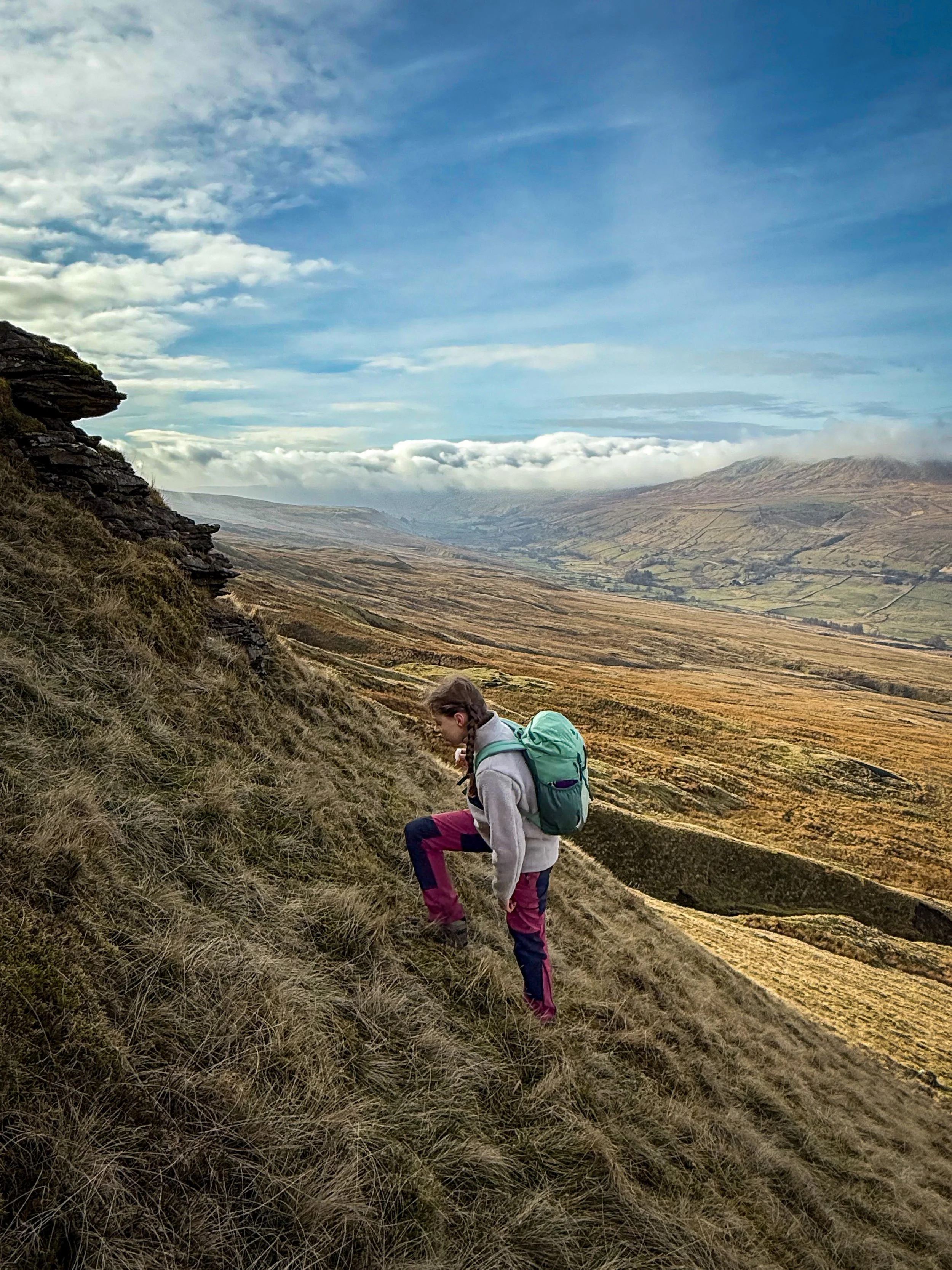 Child walking up a steep section of hillside with cloud and views to mountains and valley beyond