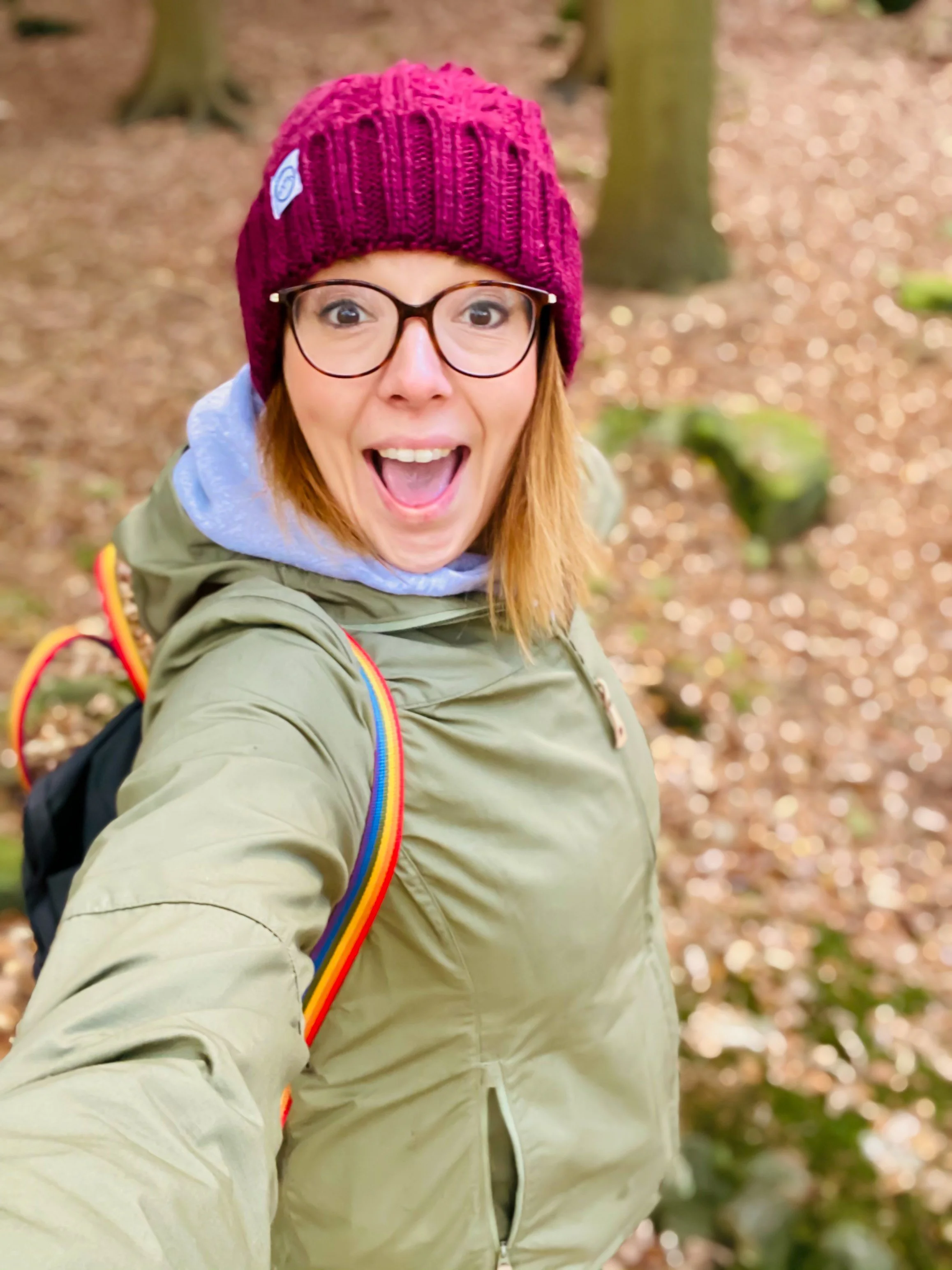 A woman with glasses and a burgundy knit beanie smiling broadly while taking a selfie outdoors in a green jacket and rainbow straps on a rucksack