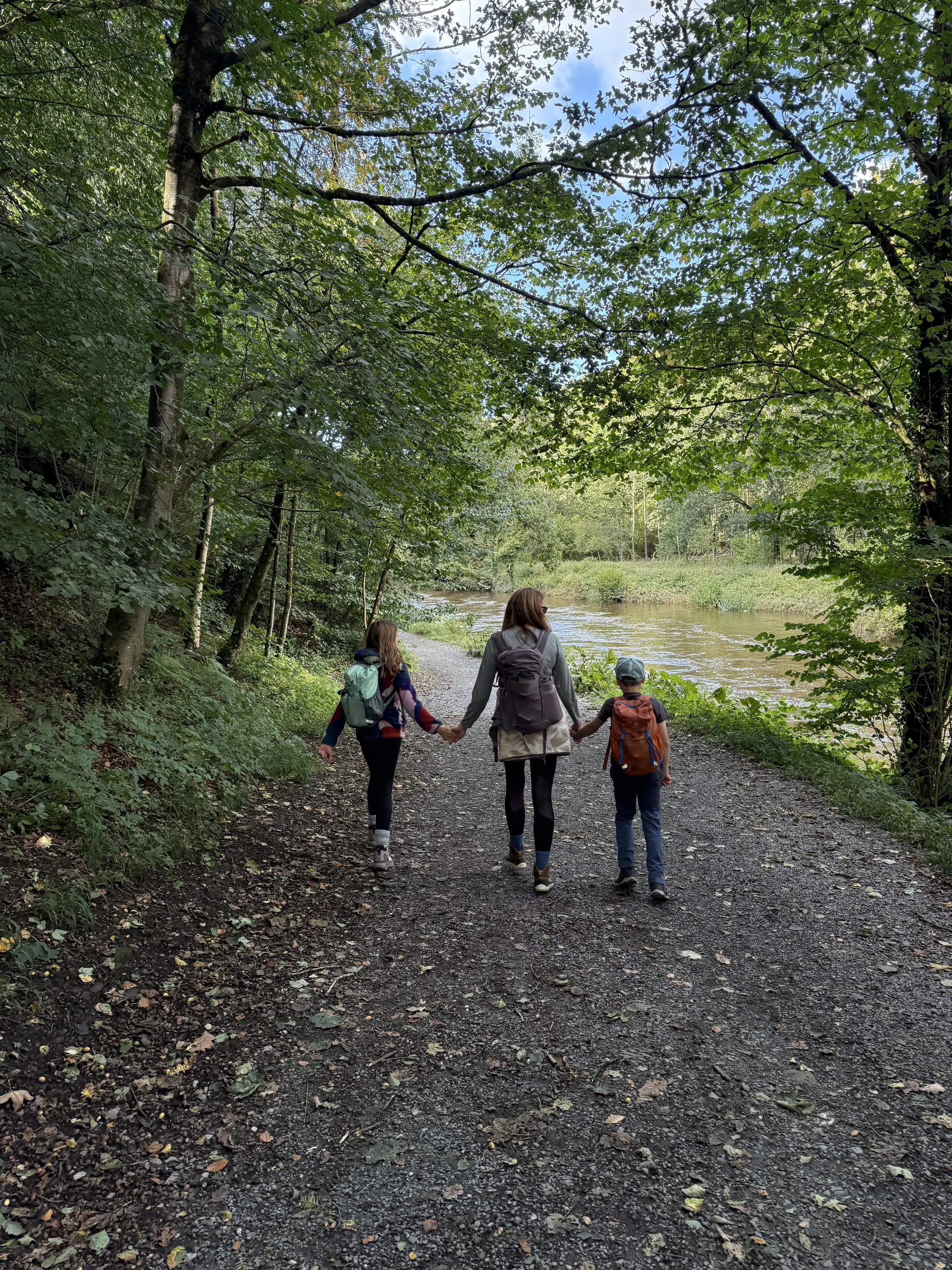 A mum walking with two kids on each side through early autumn trees on a wide path next to a river
