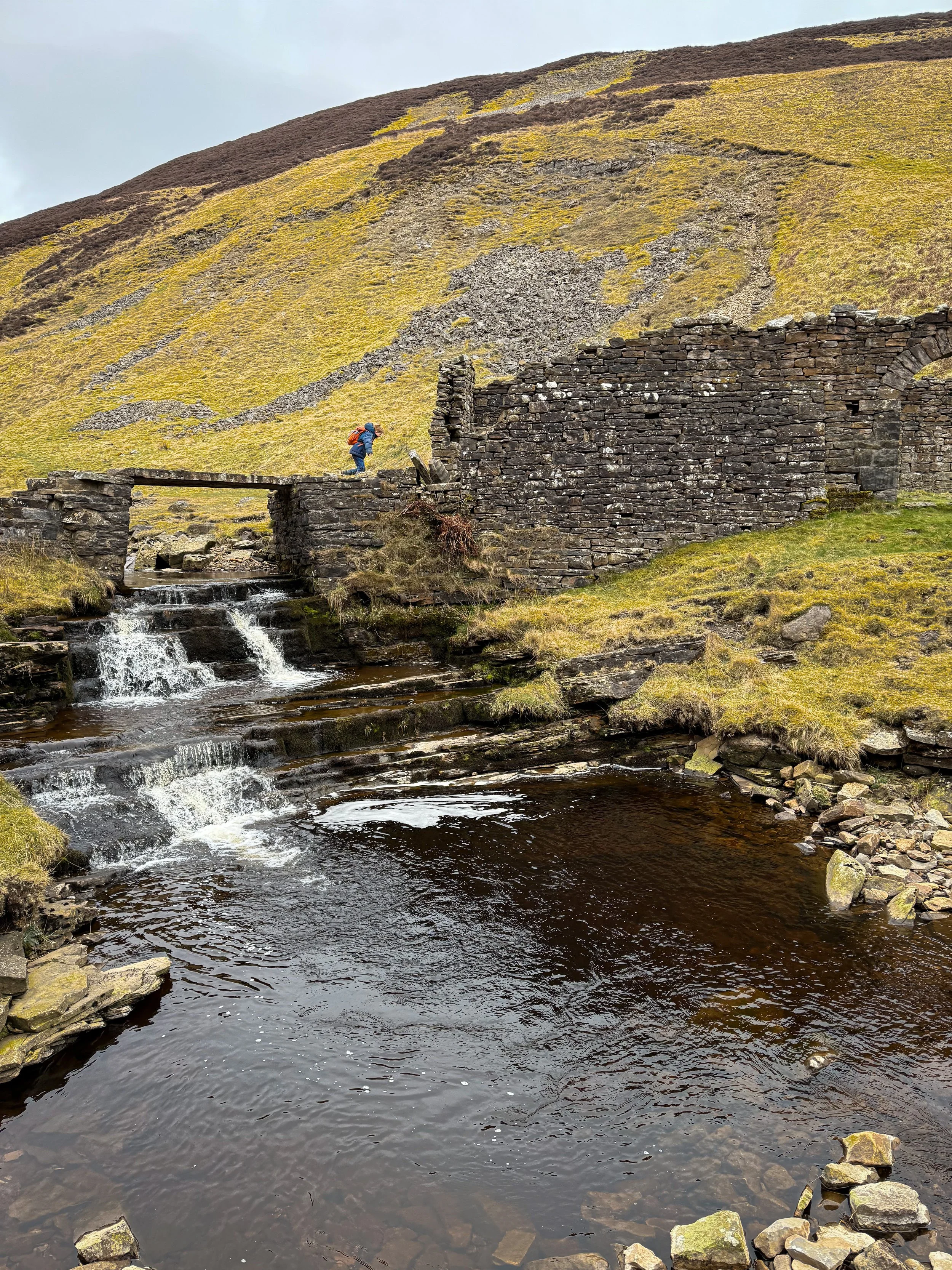 child running across a stone bridge towards a ruined building over a waterfall in the Dales