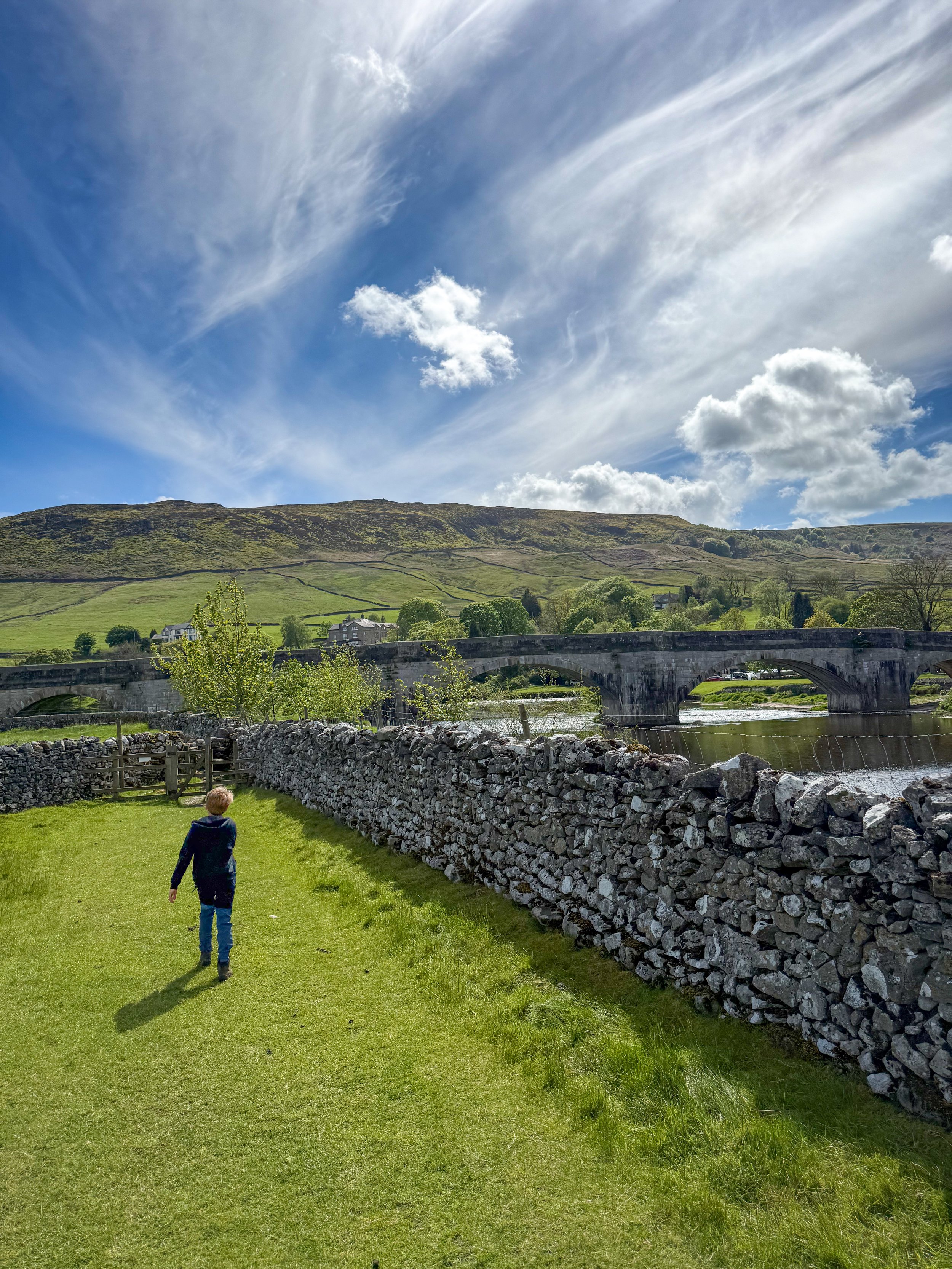 Child walking along green grass towards a bridge with fells in the distance next to a river