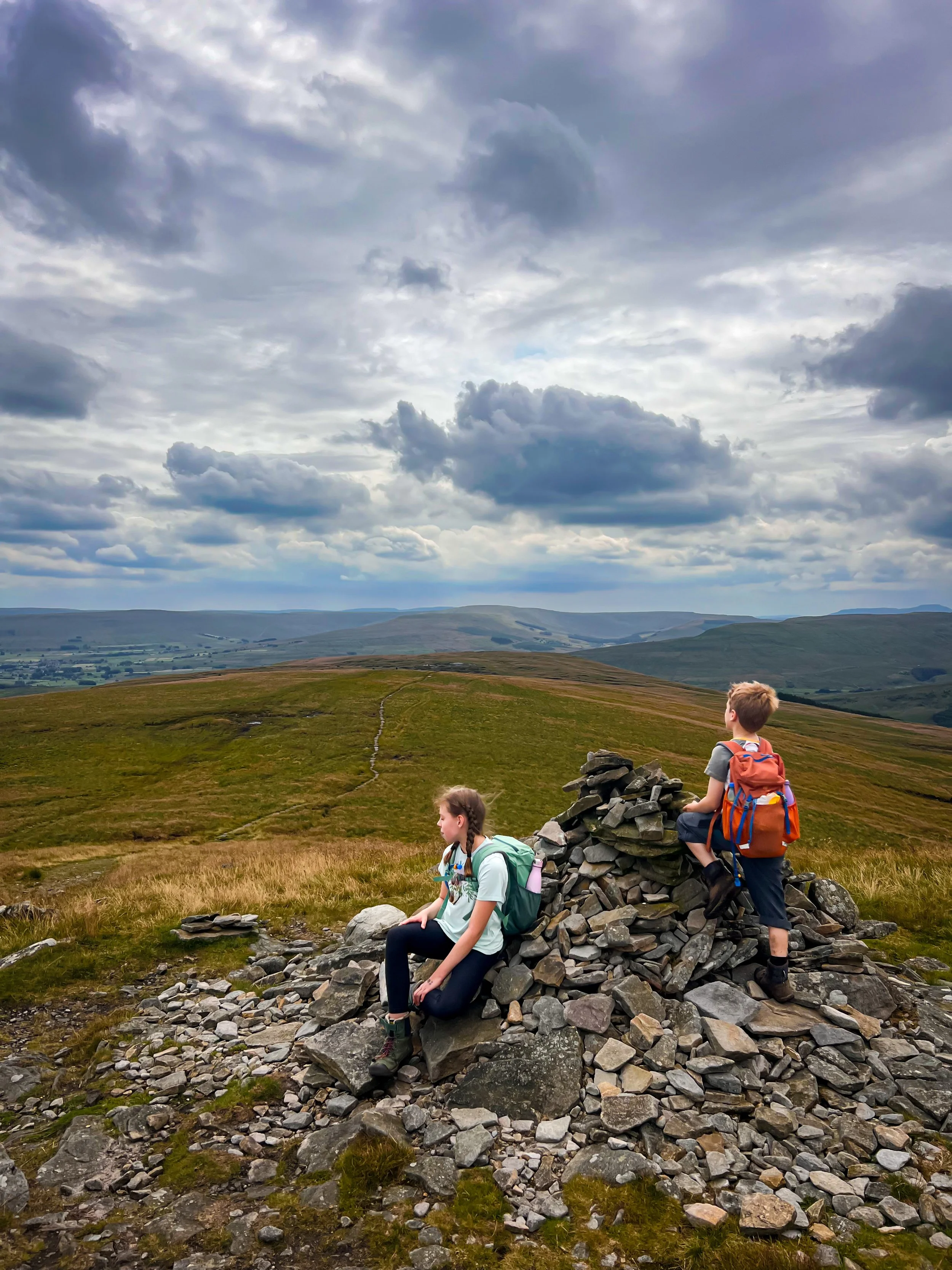 Two children on the side of a mountain with a cairn and moody grey skies