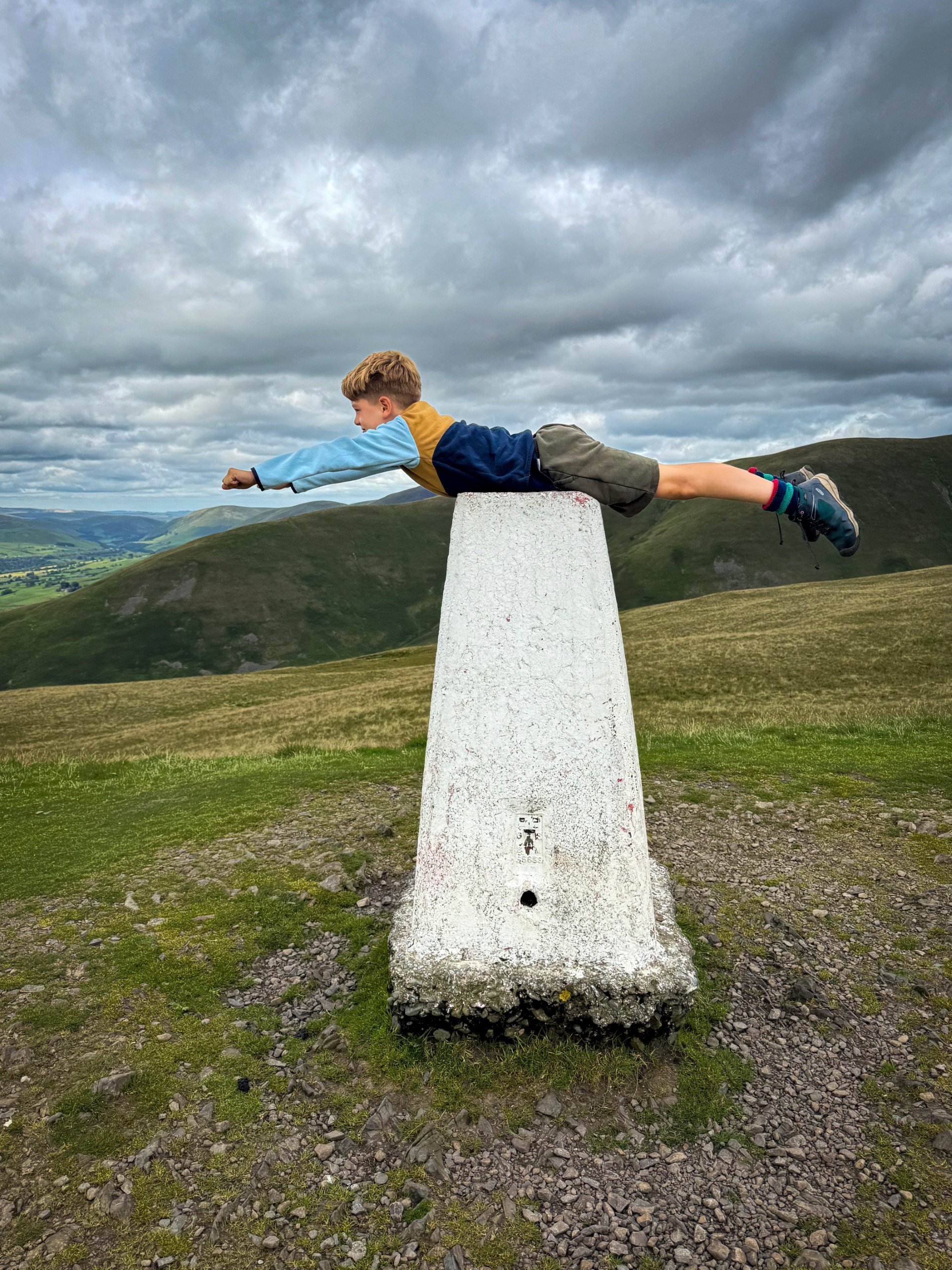Child laying on top of a trig on top of a fell with more mountains behind