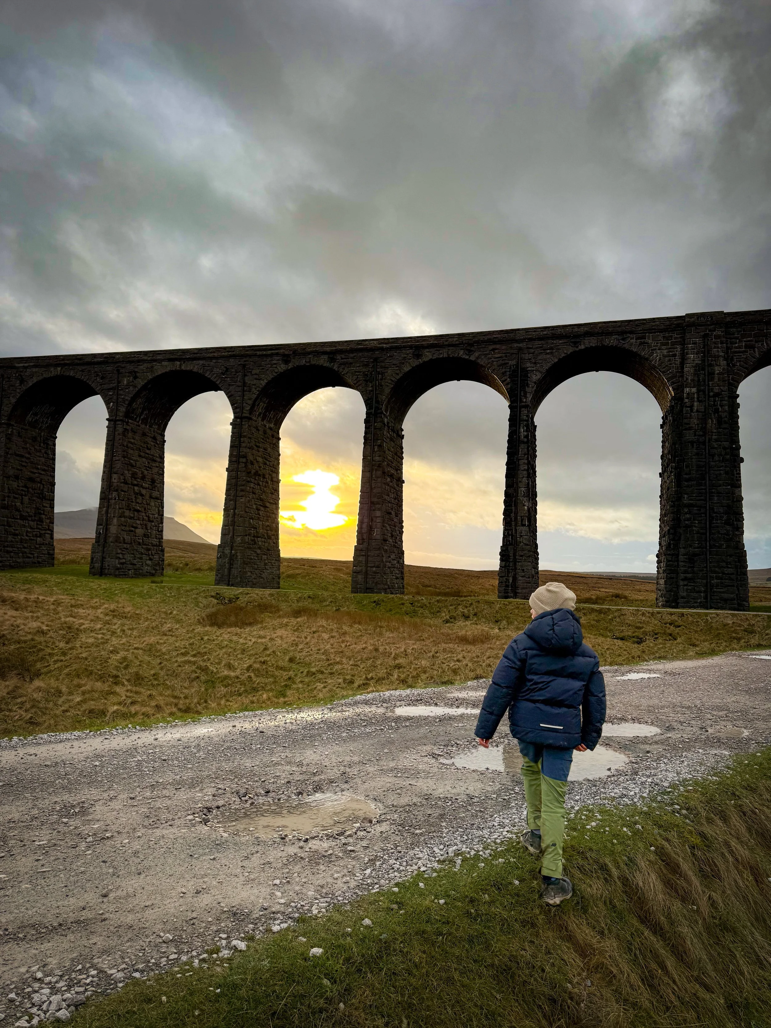 Child walking towards large viaduct arches with low winter sun in the background through the arches and grey clouds