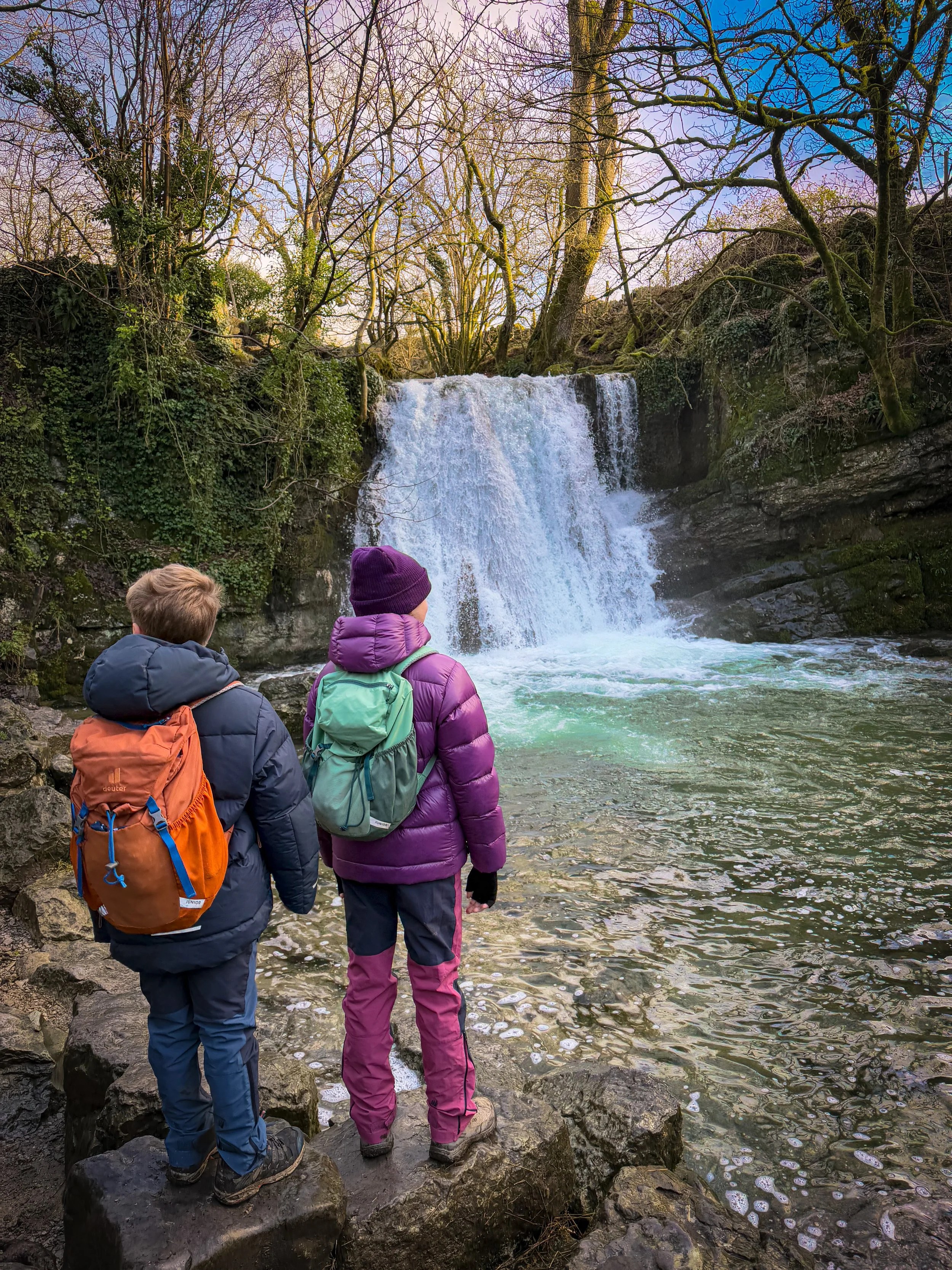 Two children standing in the winter looking at a fierce waterfall