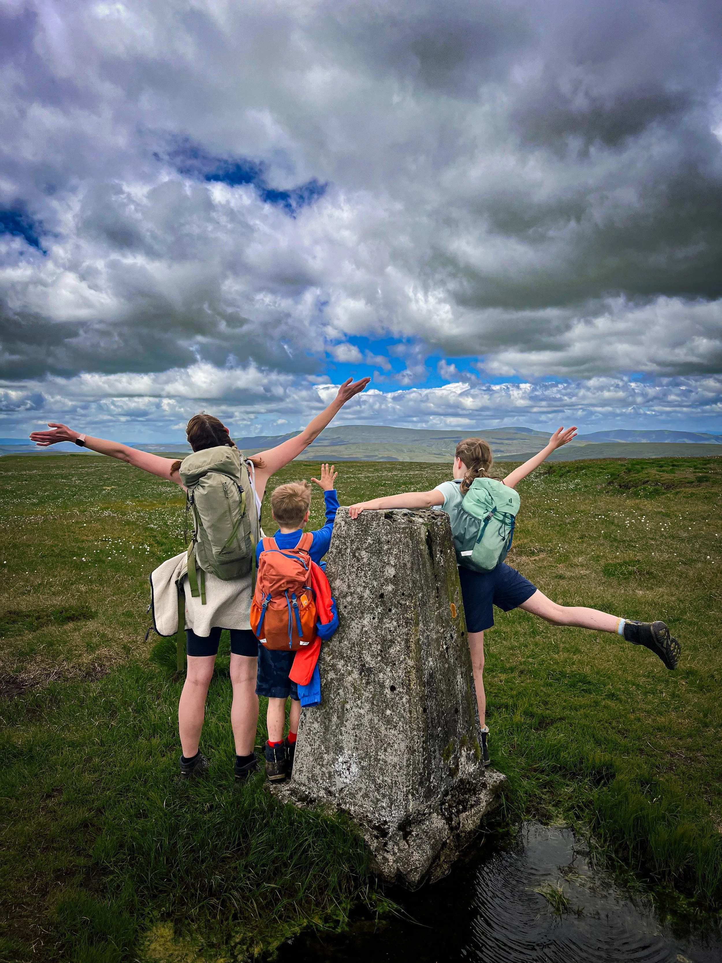 Three people standing at a trig point with arms in the air on top of a mountain with blue skies above