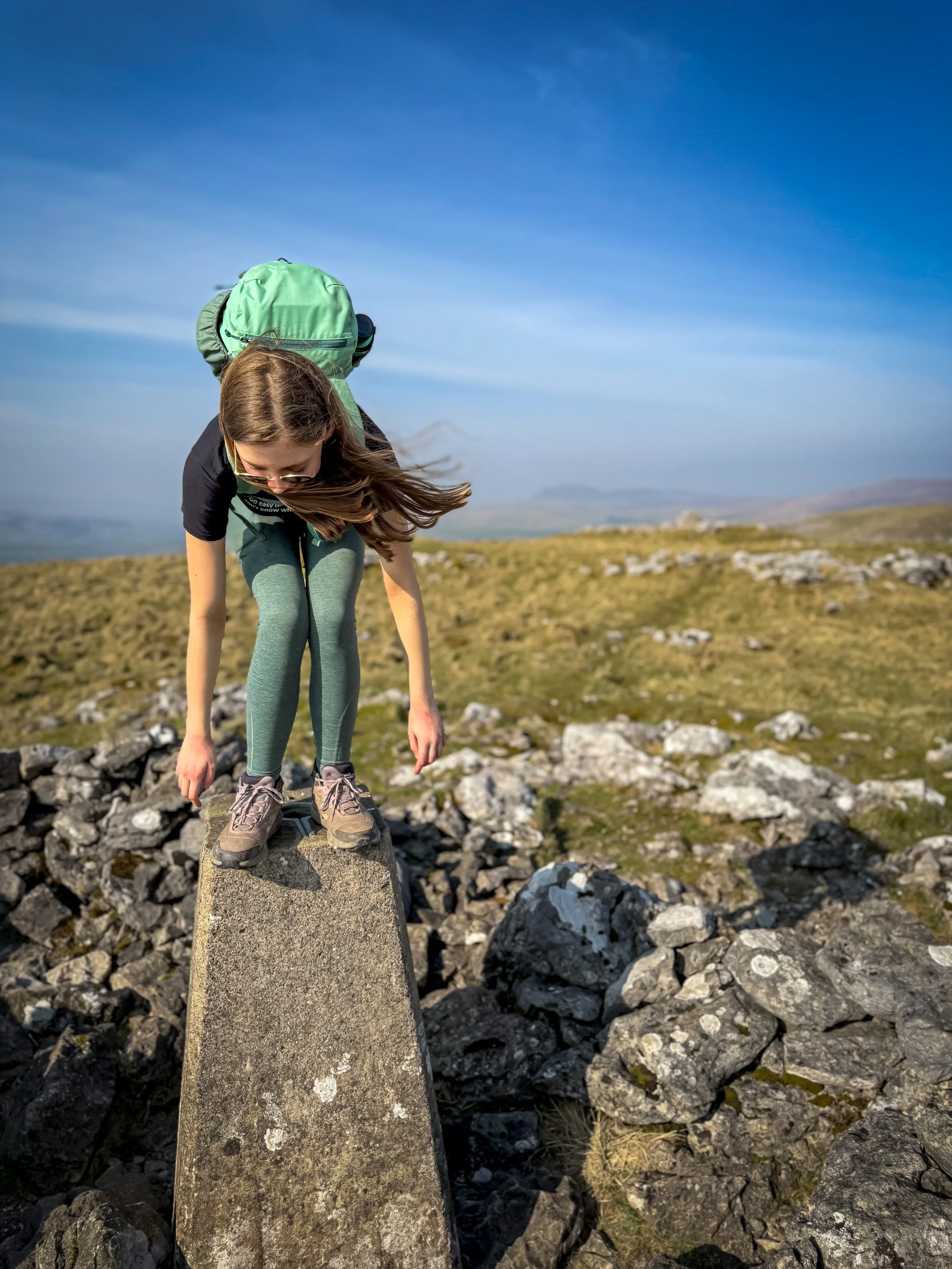 child standing on trig point, bent over with wind blowing hair gently under blue skies