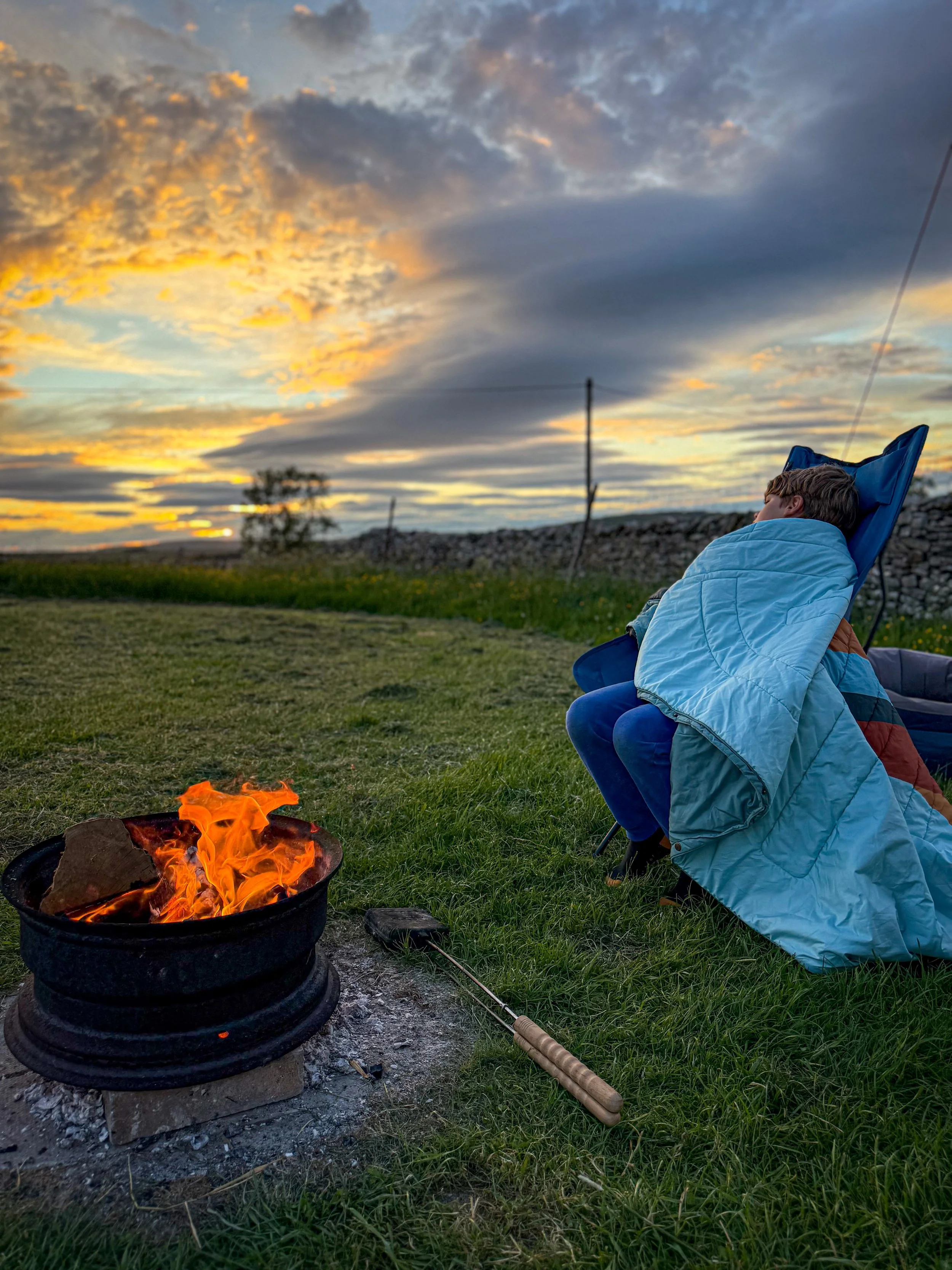 A child sat wrapped in a blanket on a camping chair next to a campfire with sunset skies and a line of dry stone wall next to mown grass