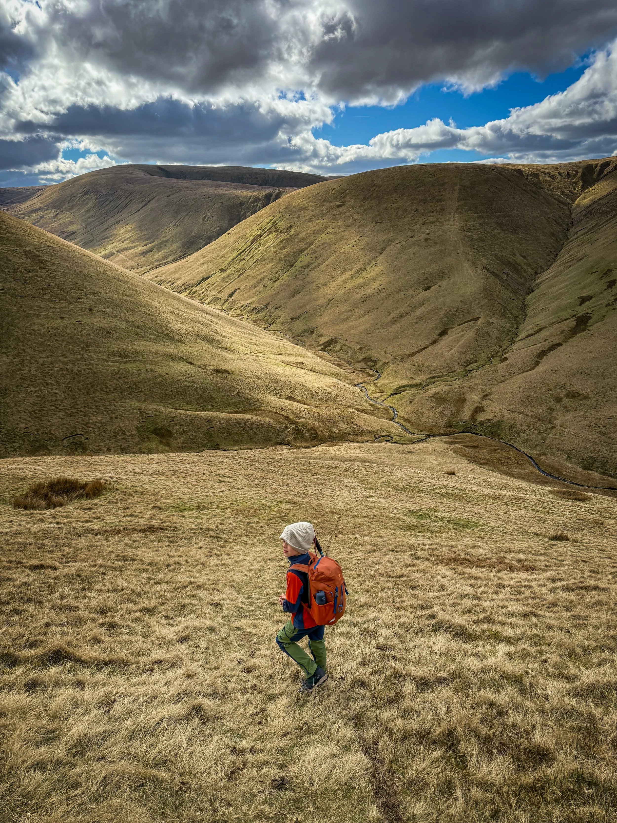 Child wearing hiking clothing and wearing  rucksack walking down a steep grassy path with grassy mountains beyond