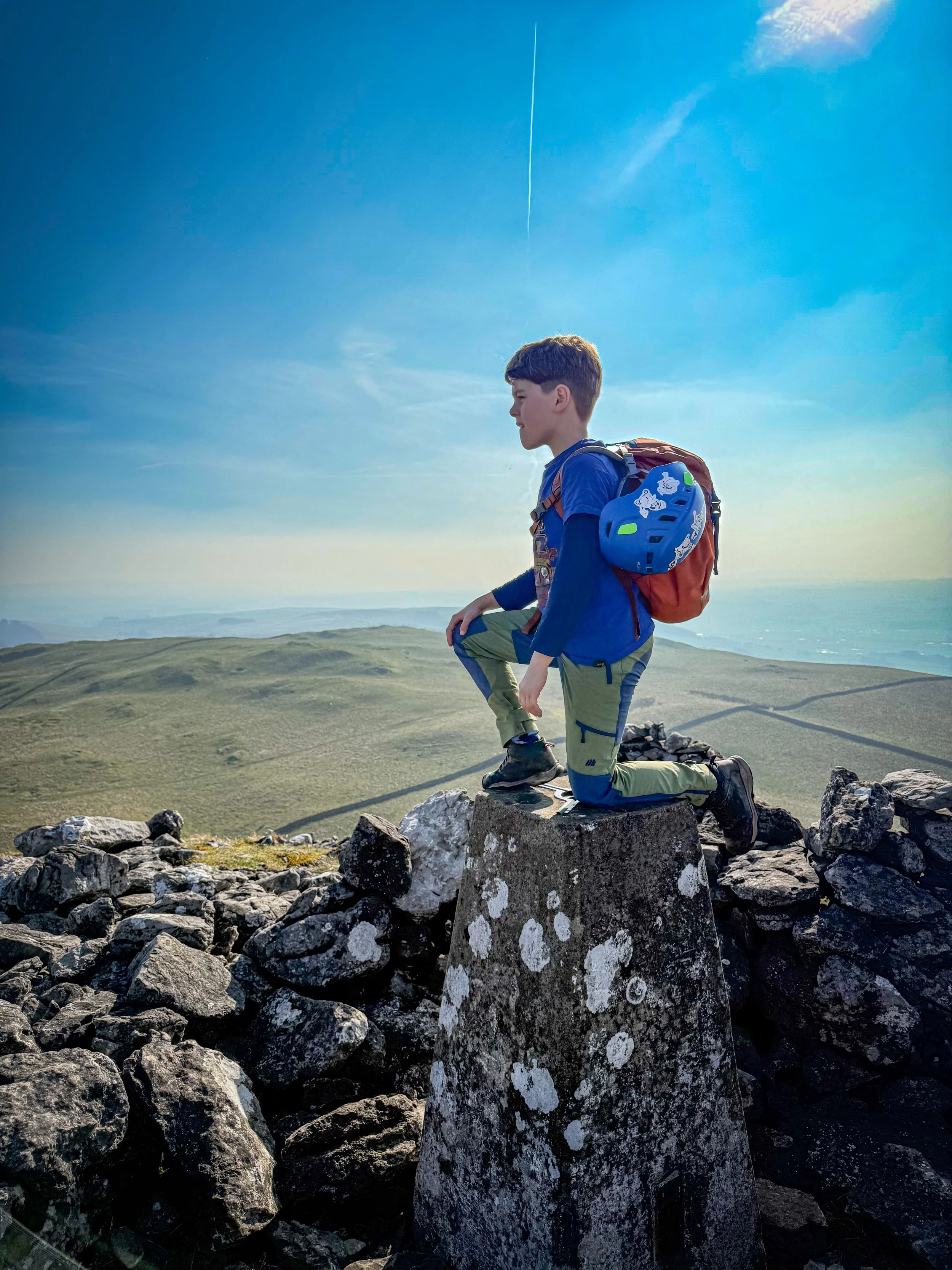 A child kneeling on one knee on top of a trig point surrounded by rocks in the distance a grassy hill he's wearing green hiking trousers and a blue tshirt with a climbing helmet on his orange backpack