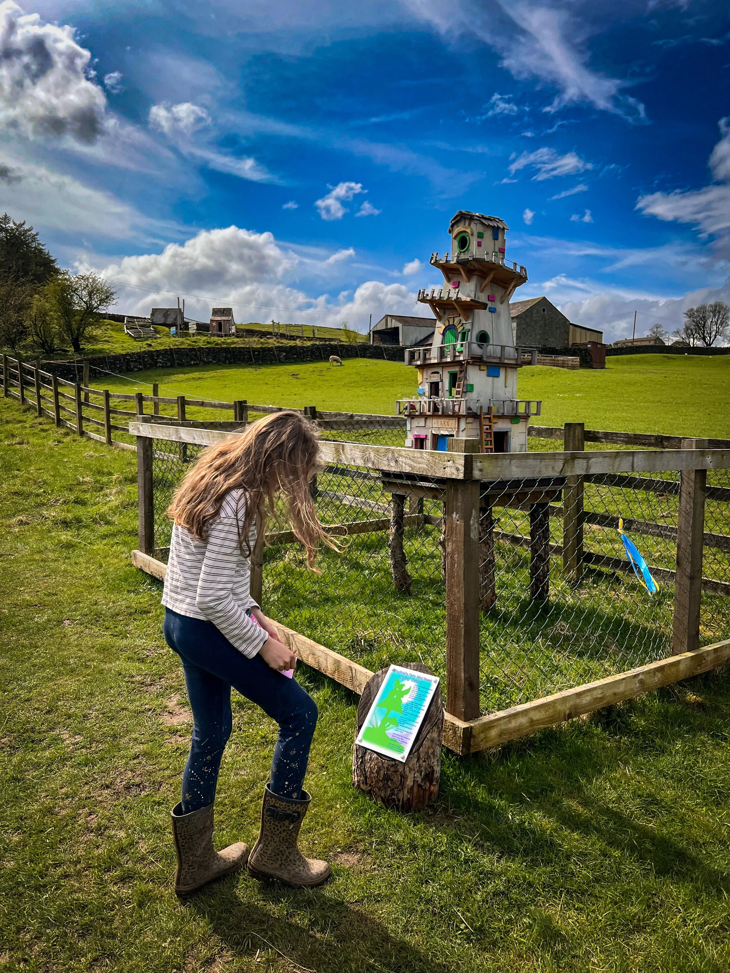 Child standing in front of a fairy house with an activity sheet on her knee and fairy information board