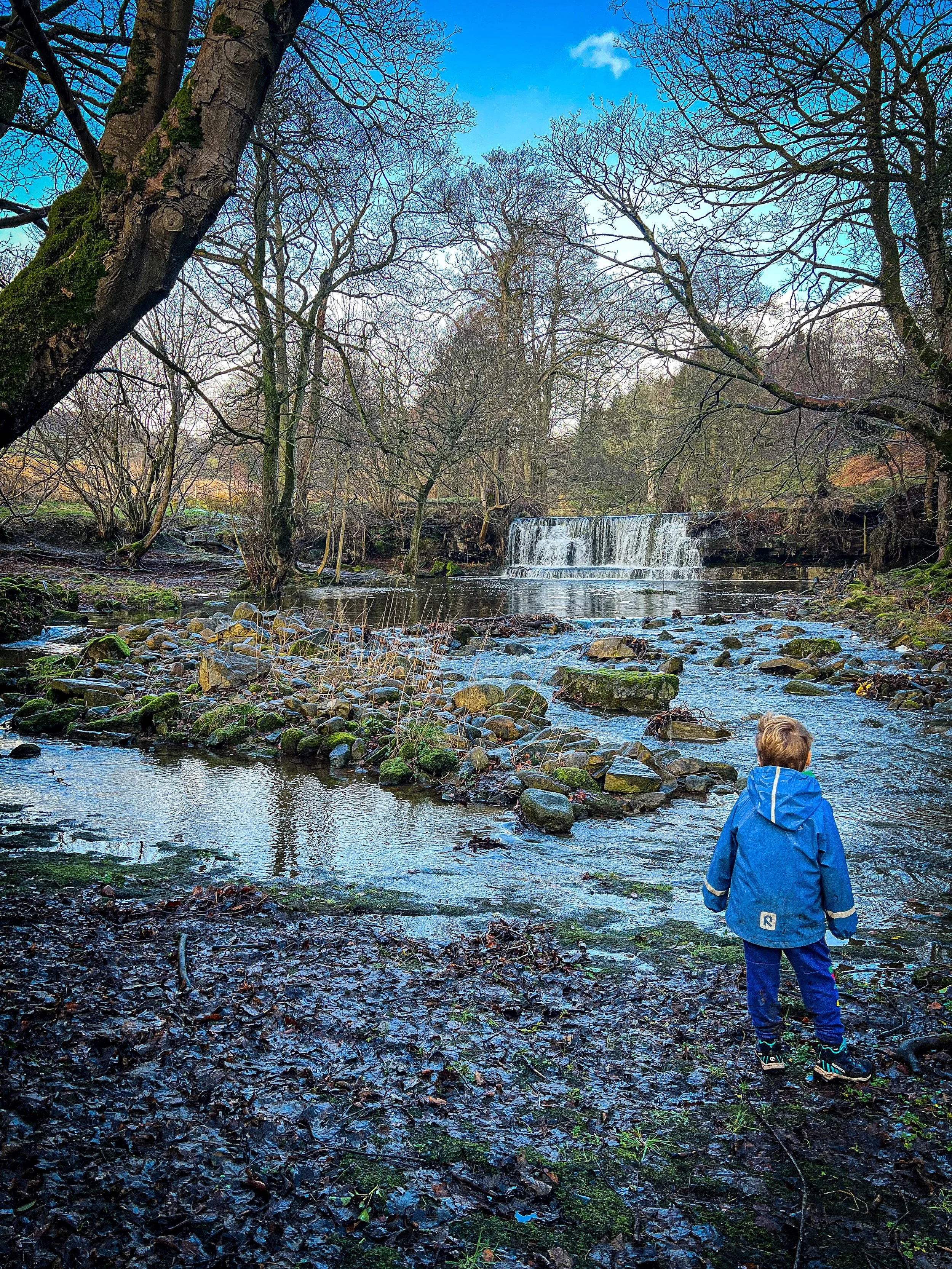 Child on a river with the waterfalls in background in winter wearing raincoat
