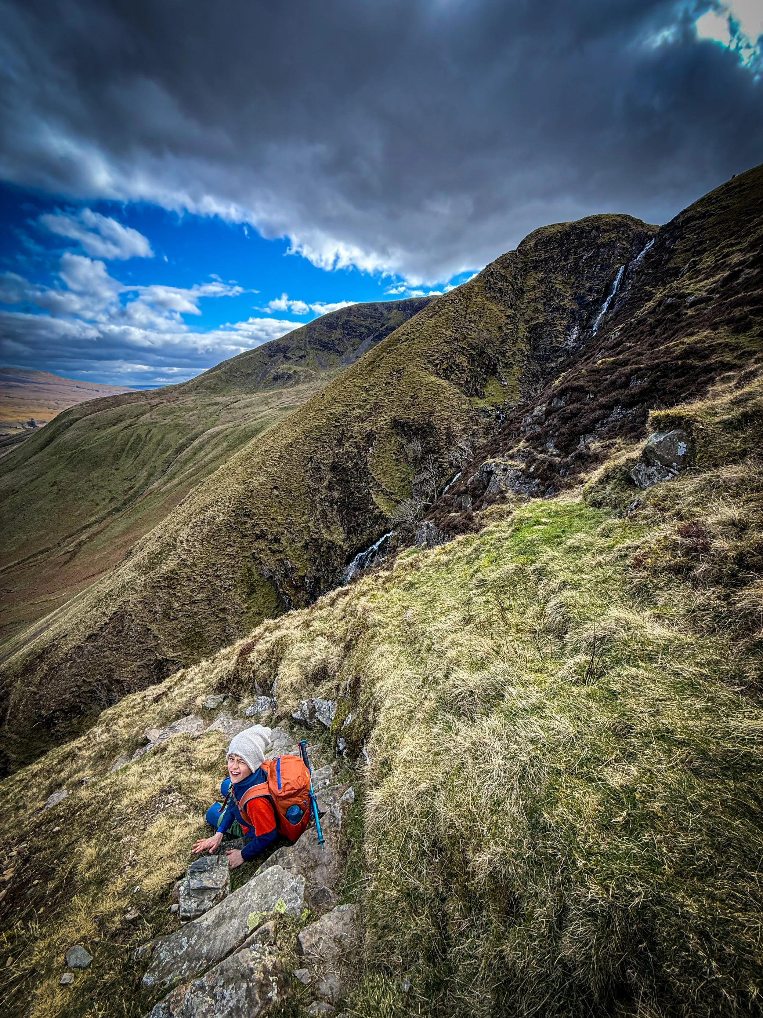 Child sat on a stony path with a waterfall in the distance and mountains beyond