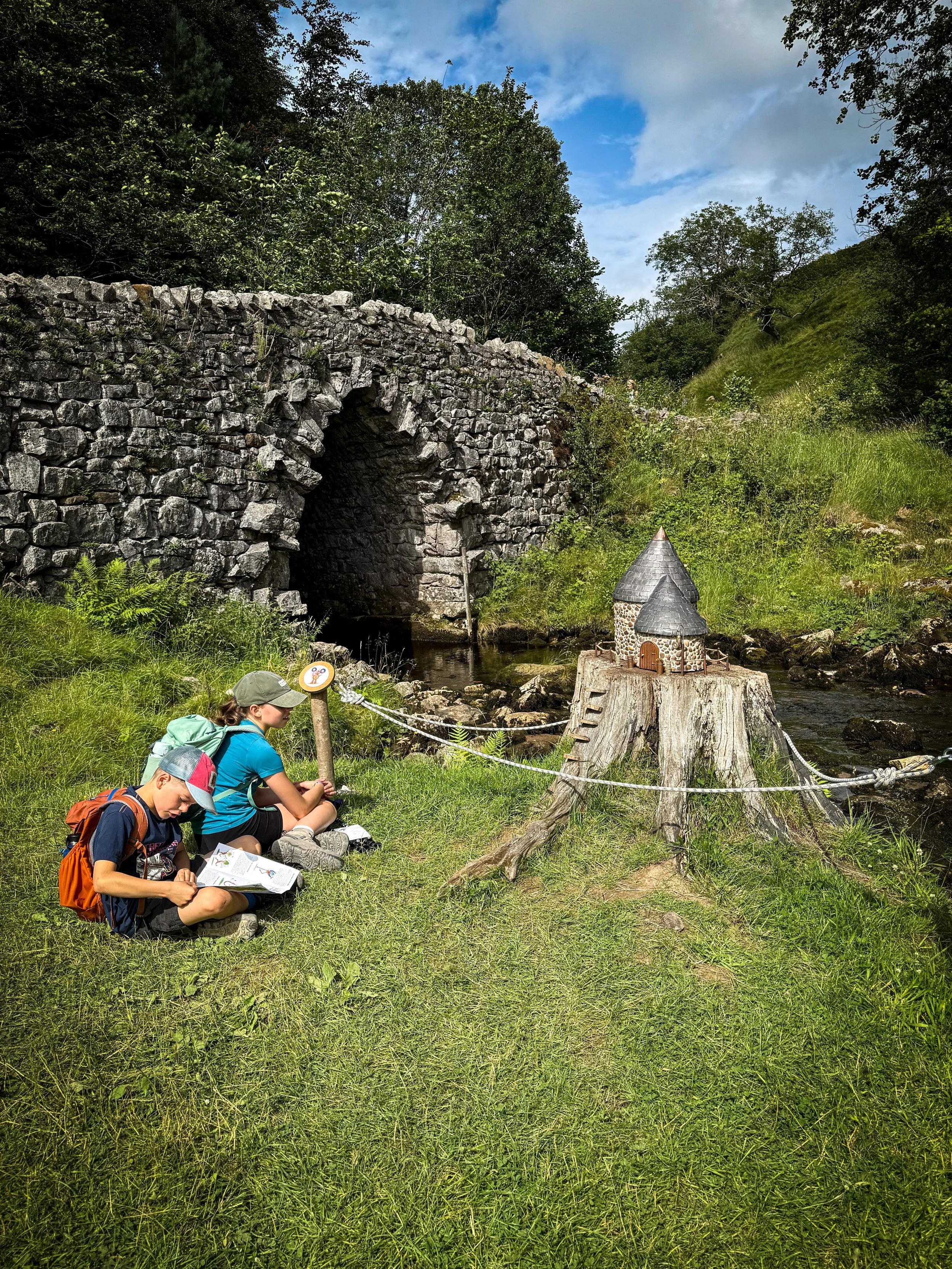 Children sitting next to a small fairy house next to a bridge