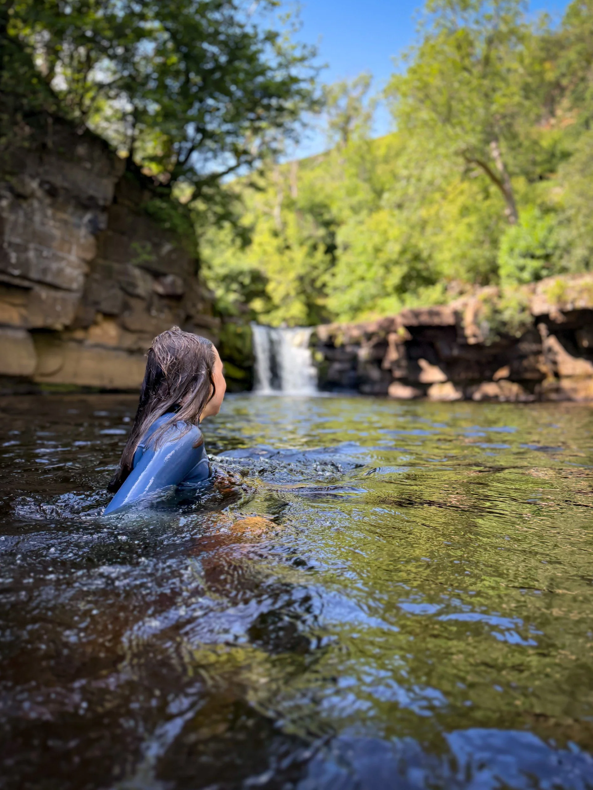 Child wearing wetsuit swimming in a waterfall in summer  with green trees and blue skies