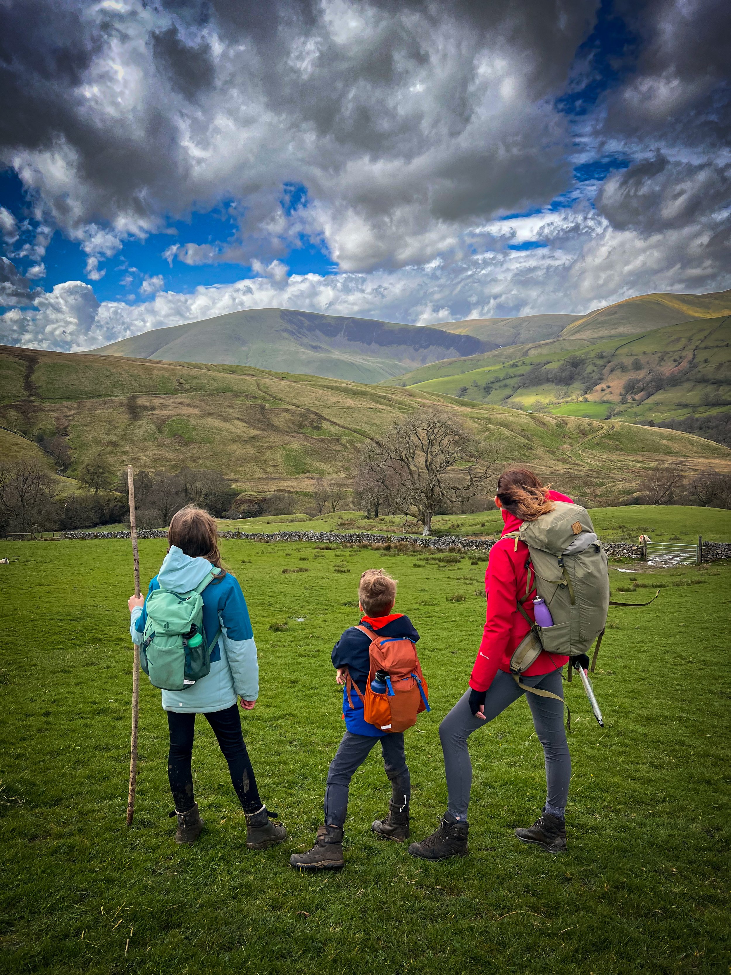 A mum and two children standing in a field all are wearing backpacks and wearing colourful coats with green rolling hills and steep mountains in head the sky is blue with fluffy clouds