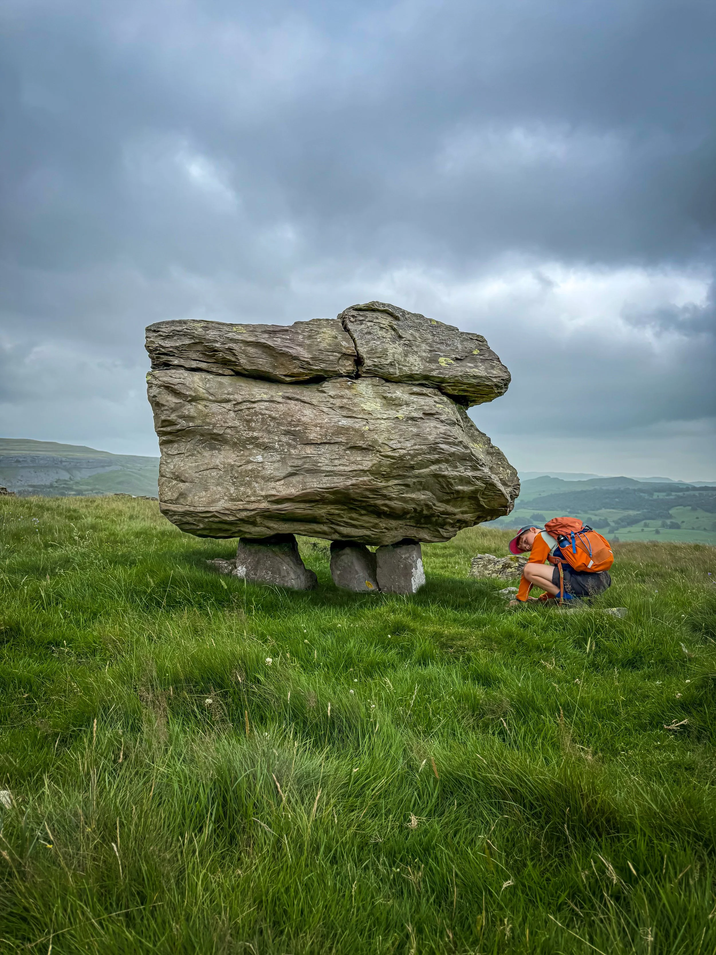 Child sat looking at a large rock perched on top of a smaller rock on top of a hill