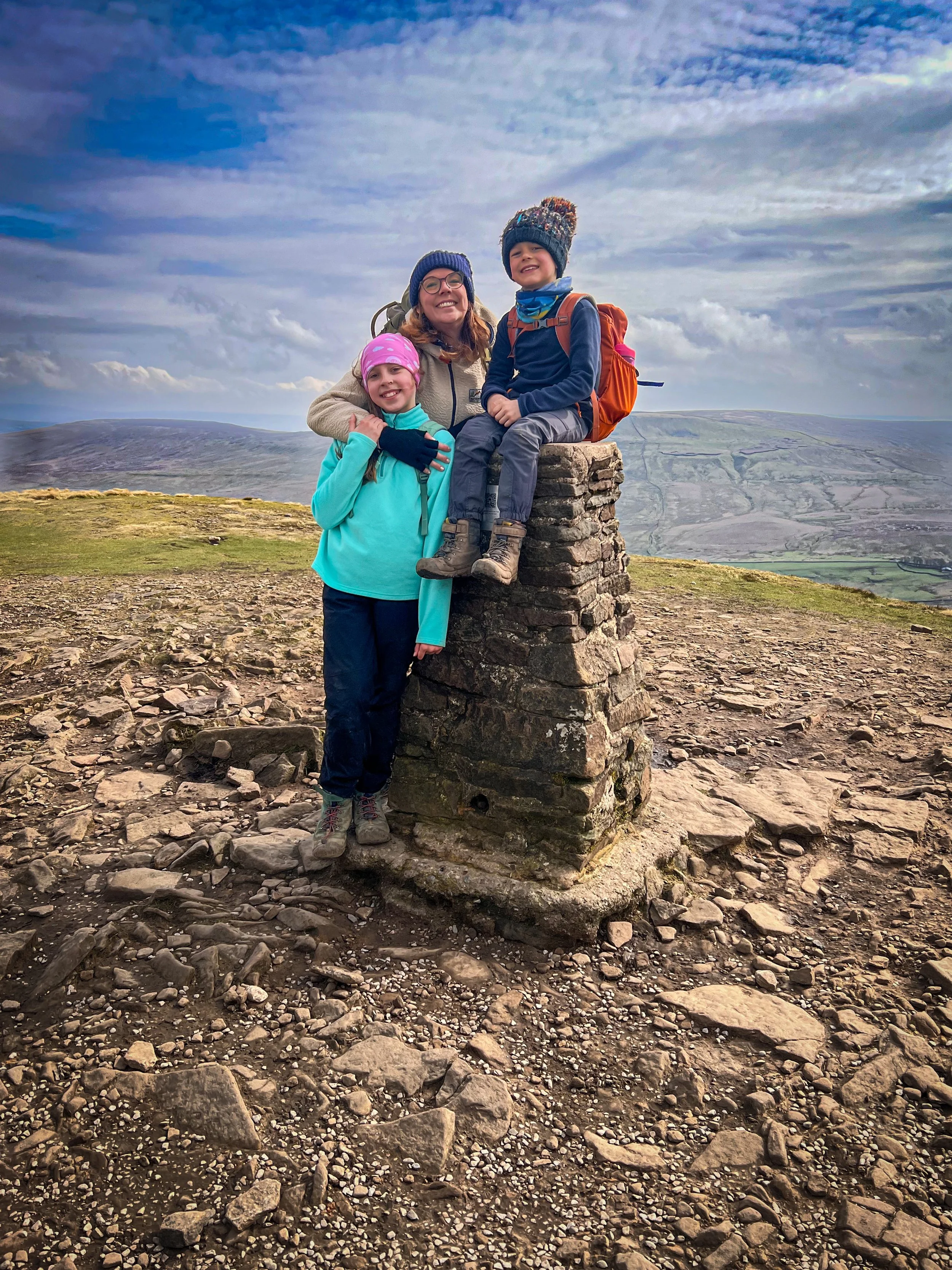 Mum and two kids standing next to the trig and sitting on the trig point of pen y ghent