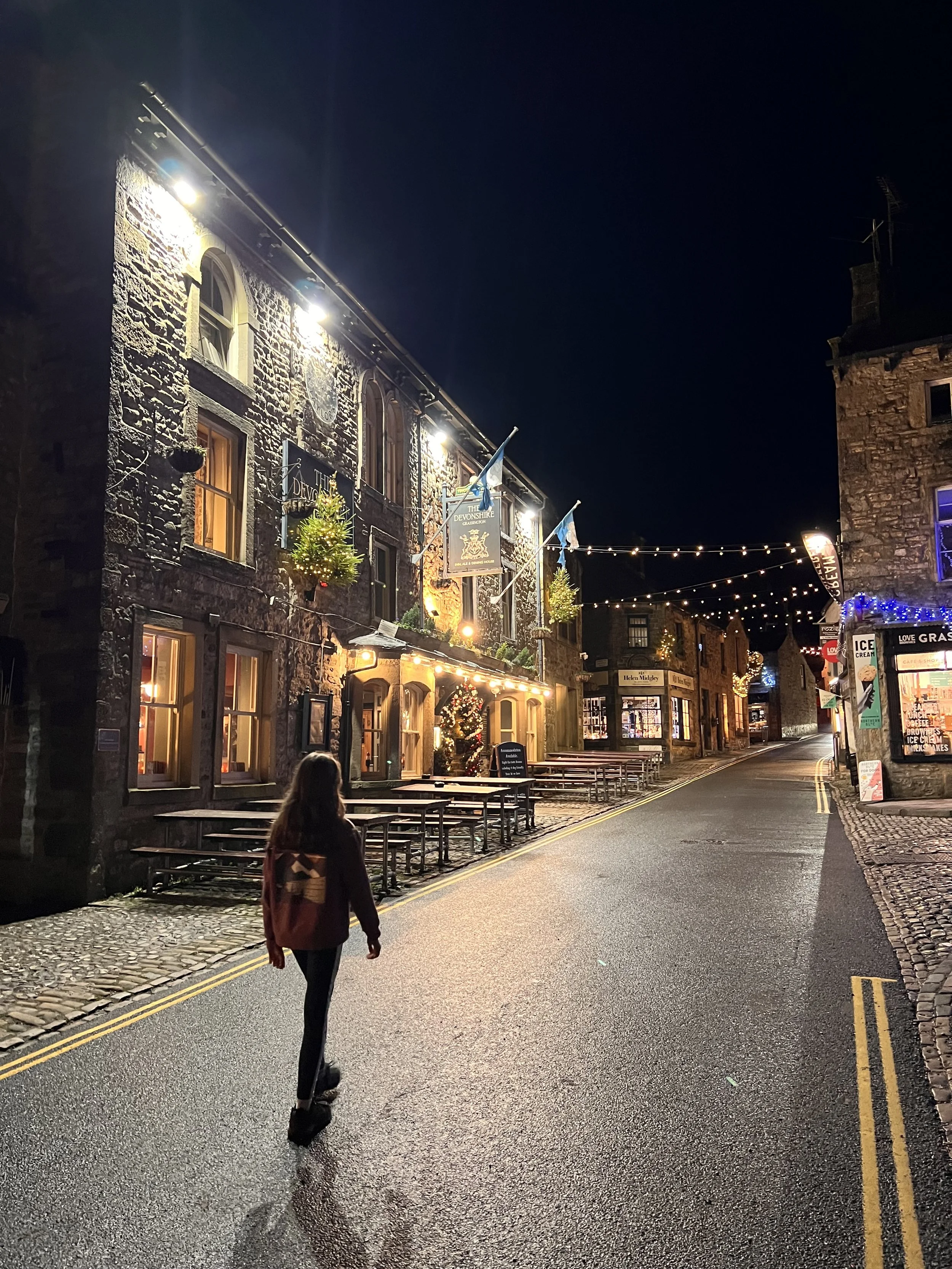 Child walking along a dark cobbled street at night next to a large building decorated for christmas to the left with light pooling on the ground