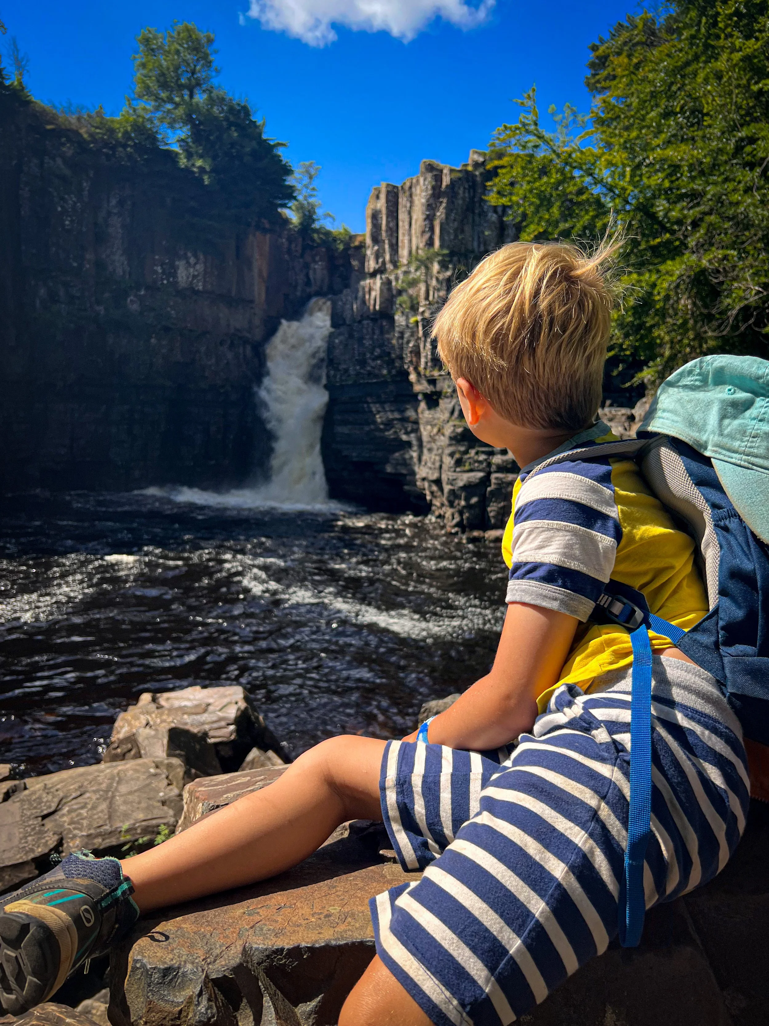 Child sat on a rock in front of a huge raging waterfall and plunge pool in the summer under blue sky