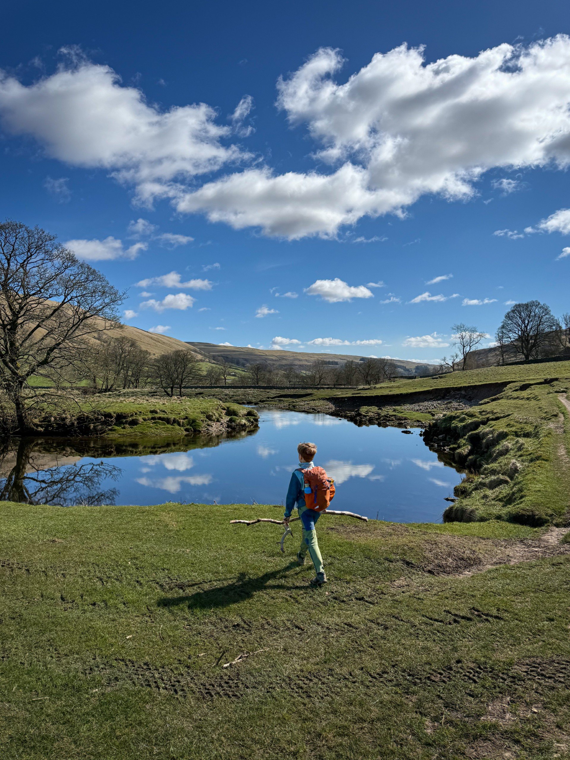 Child walking with large stick looking at calm river under a blue sky in the countryside