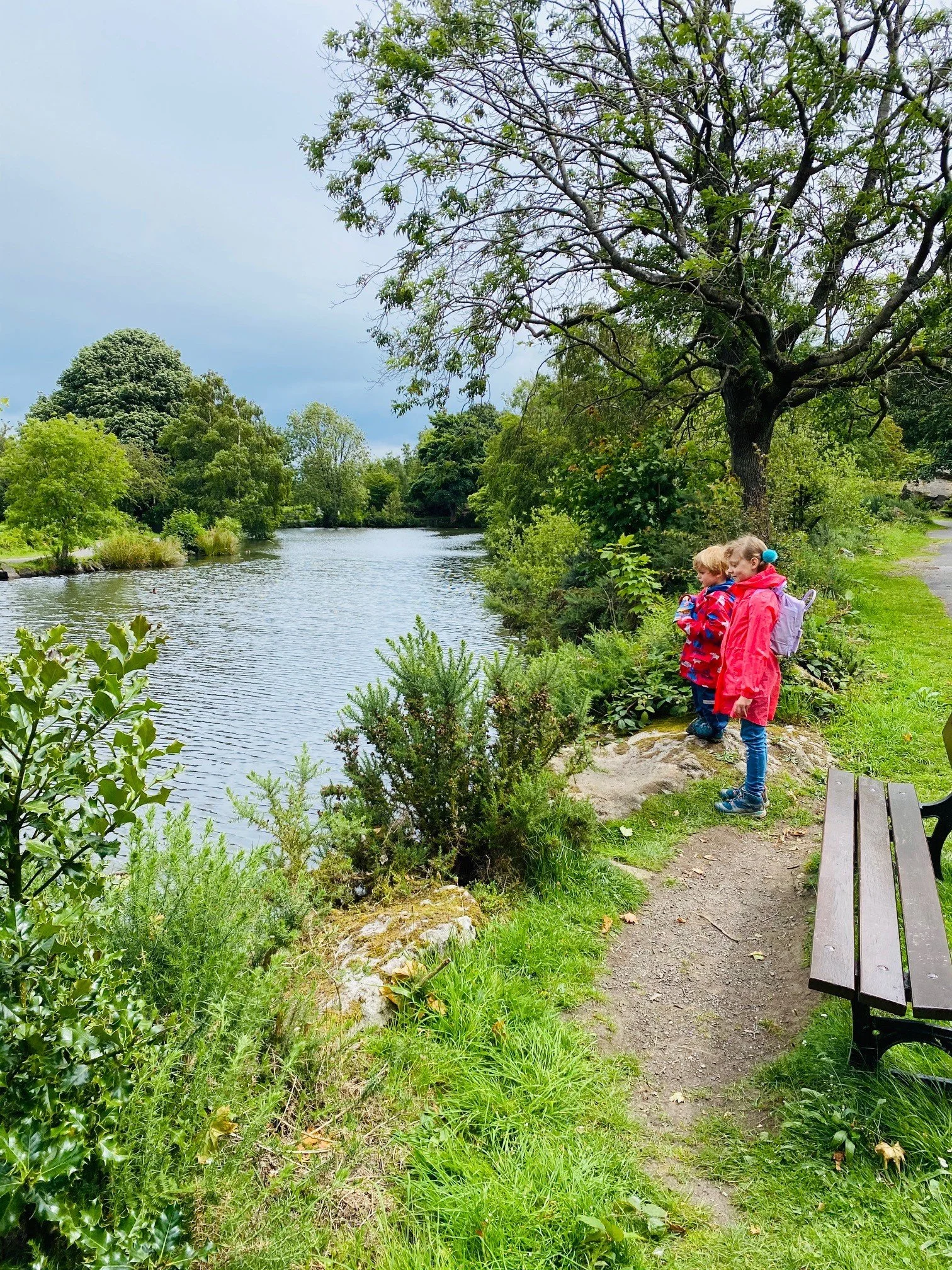 Two children standing next to a bench with Ilkley Tarn to their left and trees surrounding