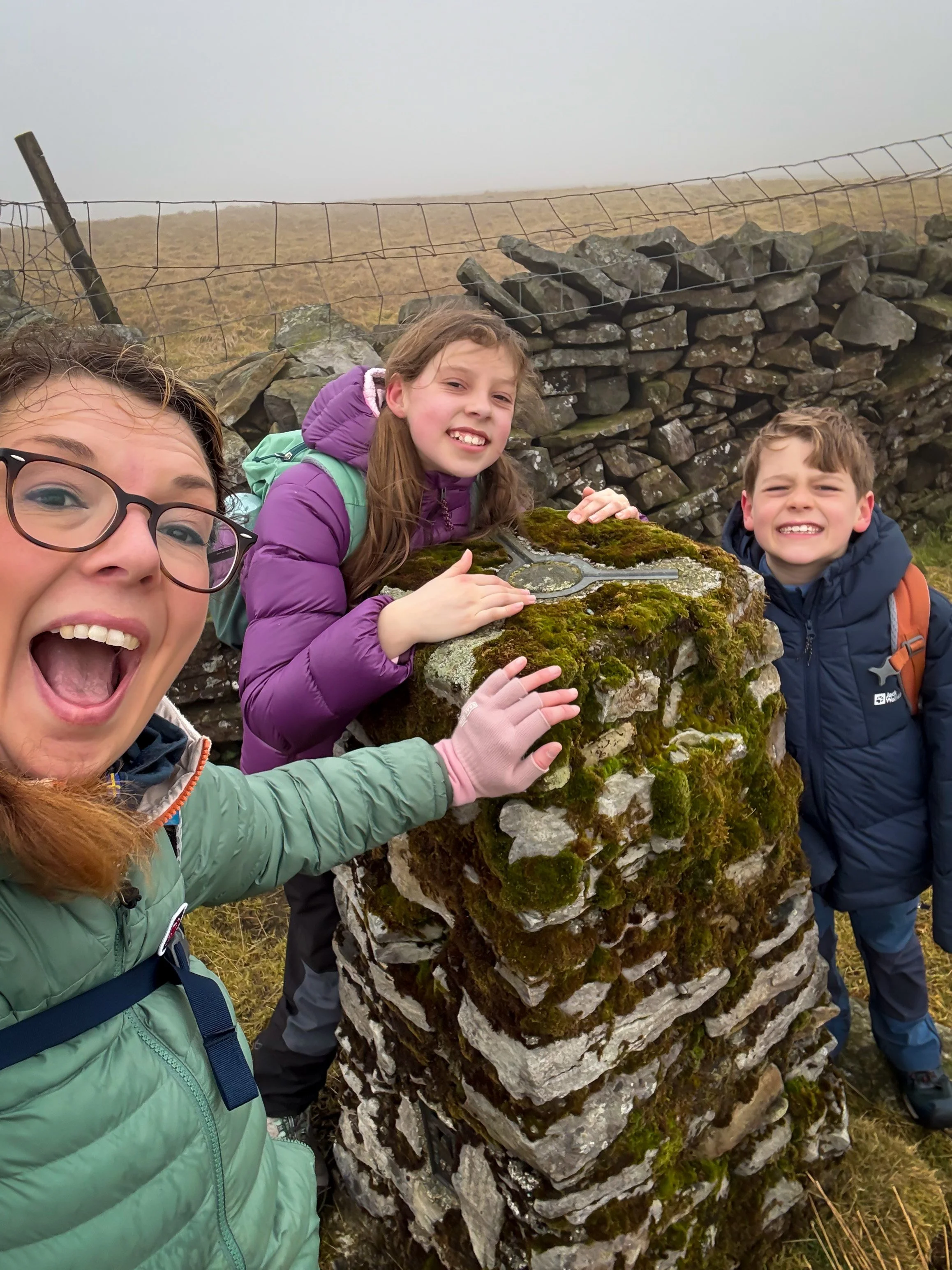 An adult and two children stood next to a stone built trig point holding the top with a wall and cloud behind
