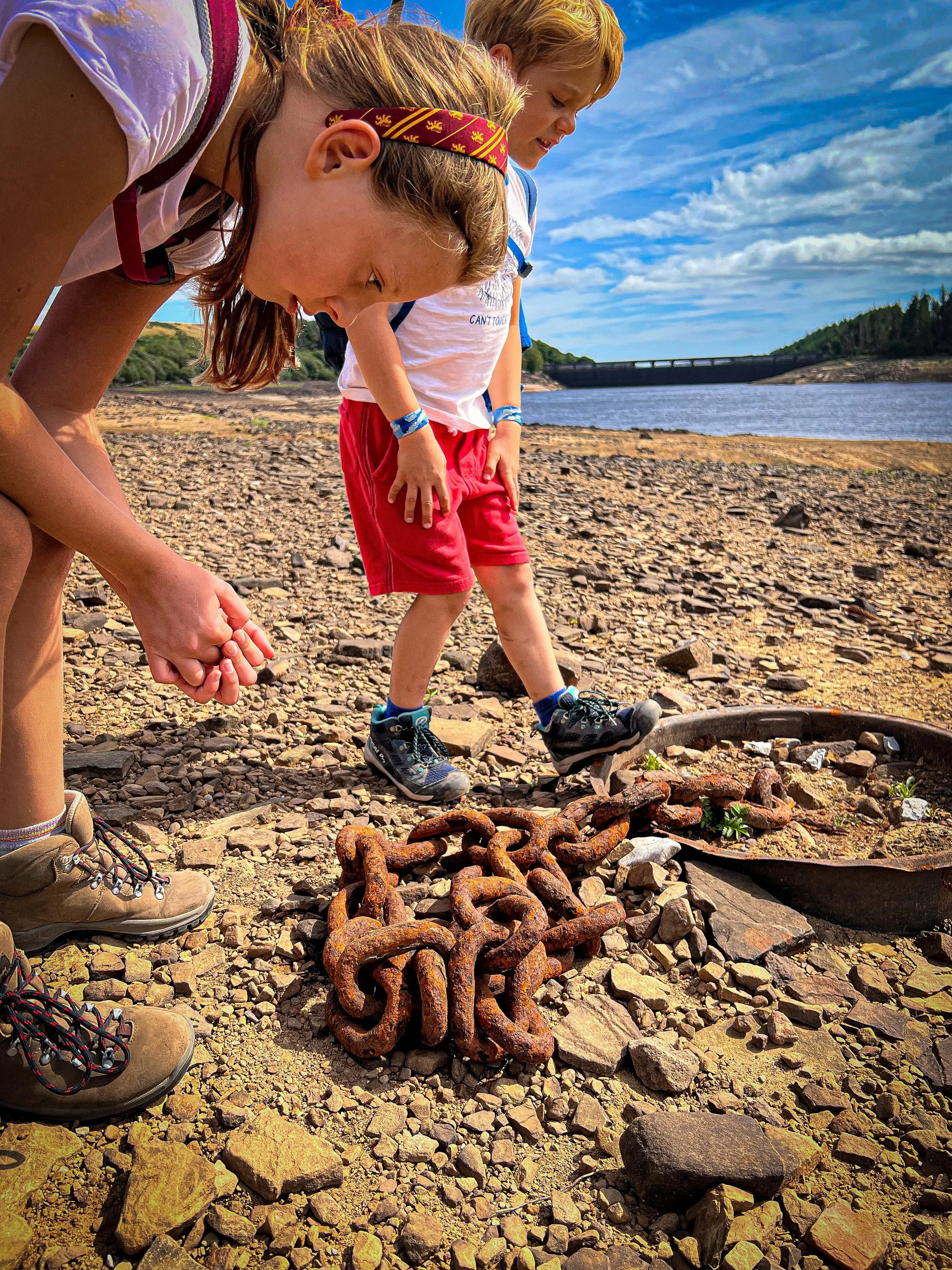 Child looking at a rusty chain on the dry bed of a reservoir with the reservoir and dam wall in the background