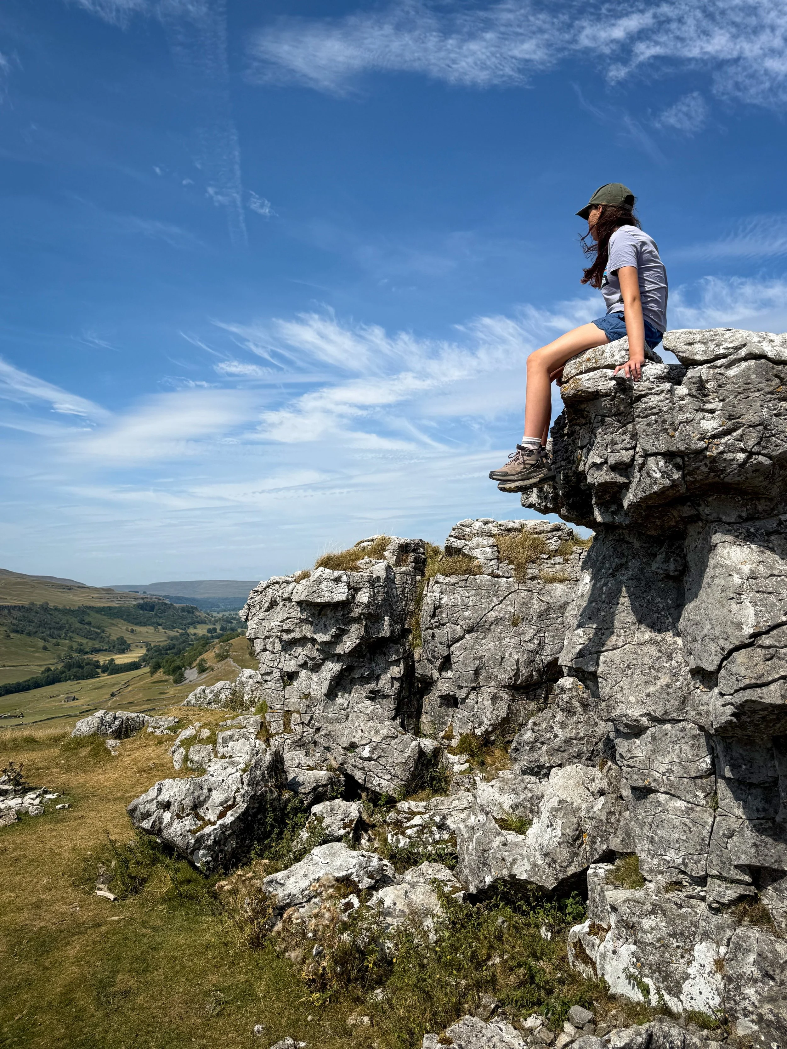 child sat on a limestone rocky outcrop with views beyond to dales and grassy hills in summer under blue sky