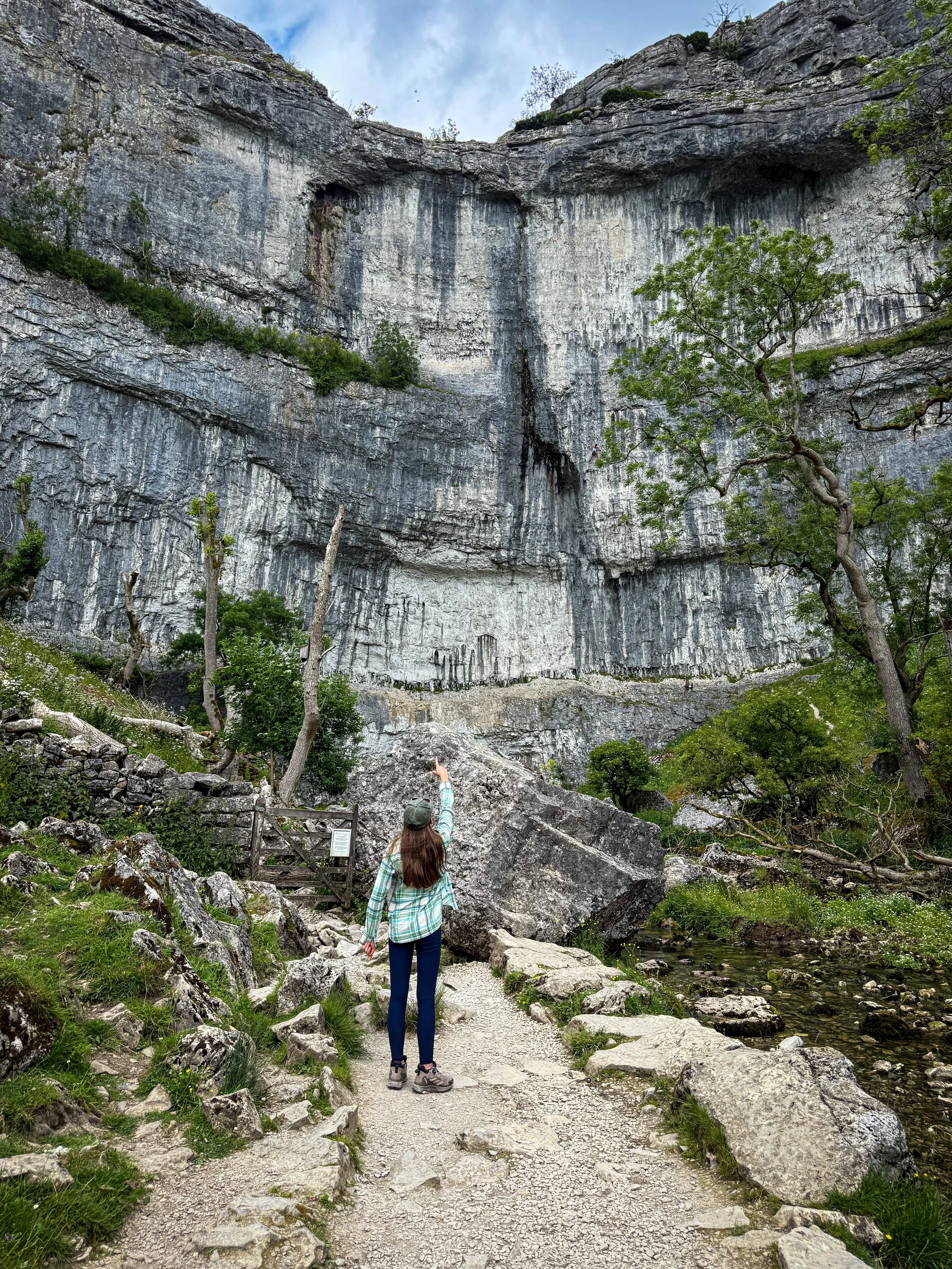 Child standing and pointing up at Malham Cove the rocky cliff face towering over her with a dry stone wall ahead