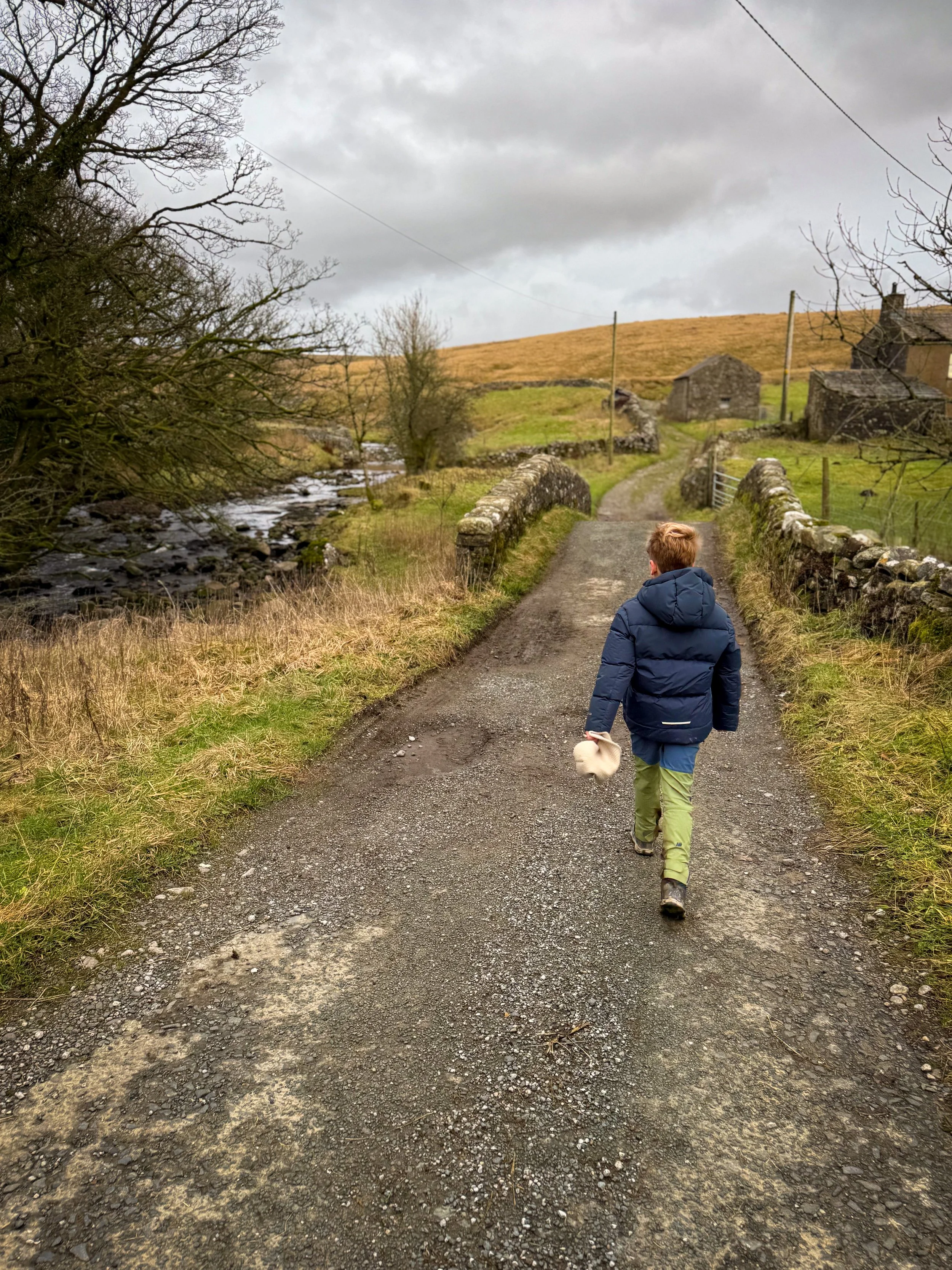 Child walking over small stone bridge with farm in the background