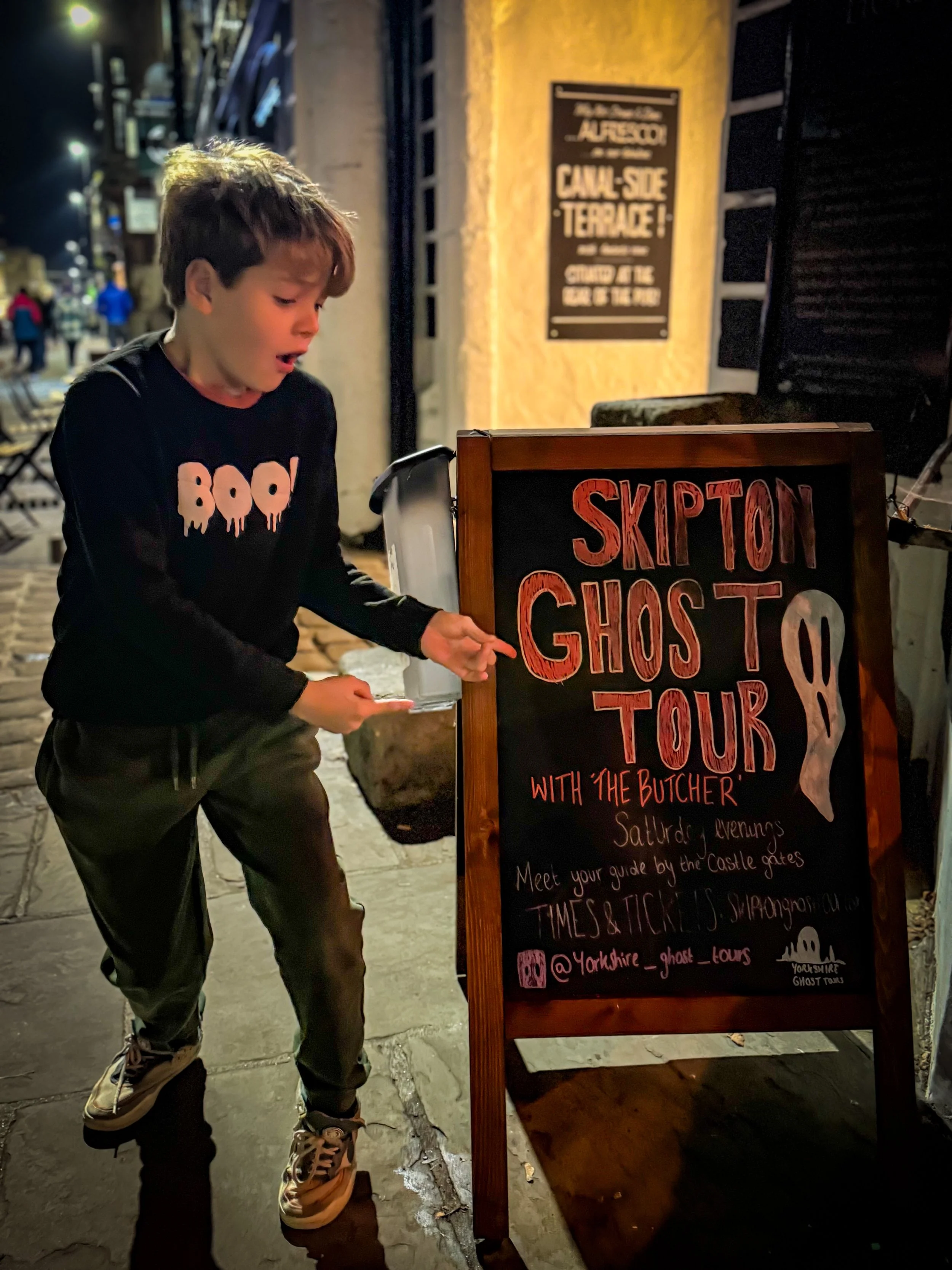 Child wearing Halloween tshirt pointing at a sign for a ghost tour