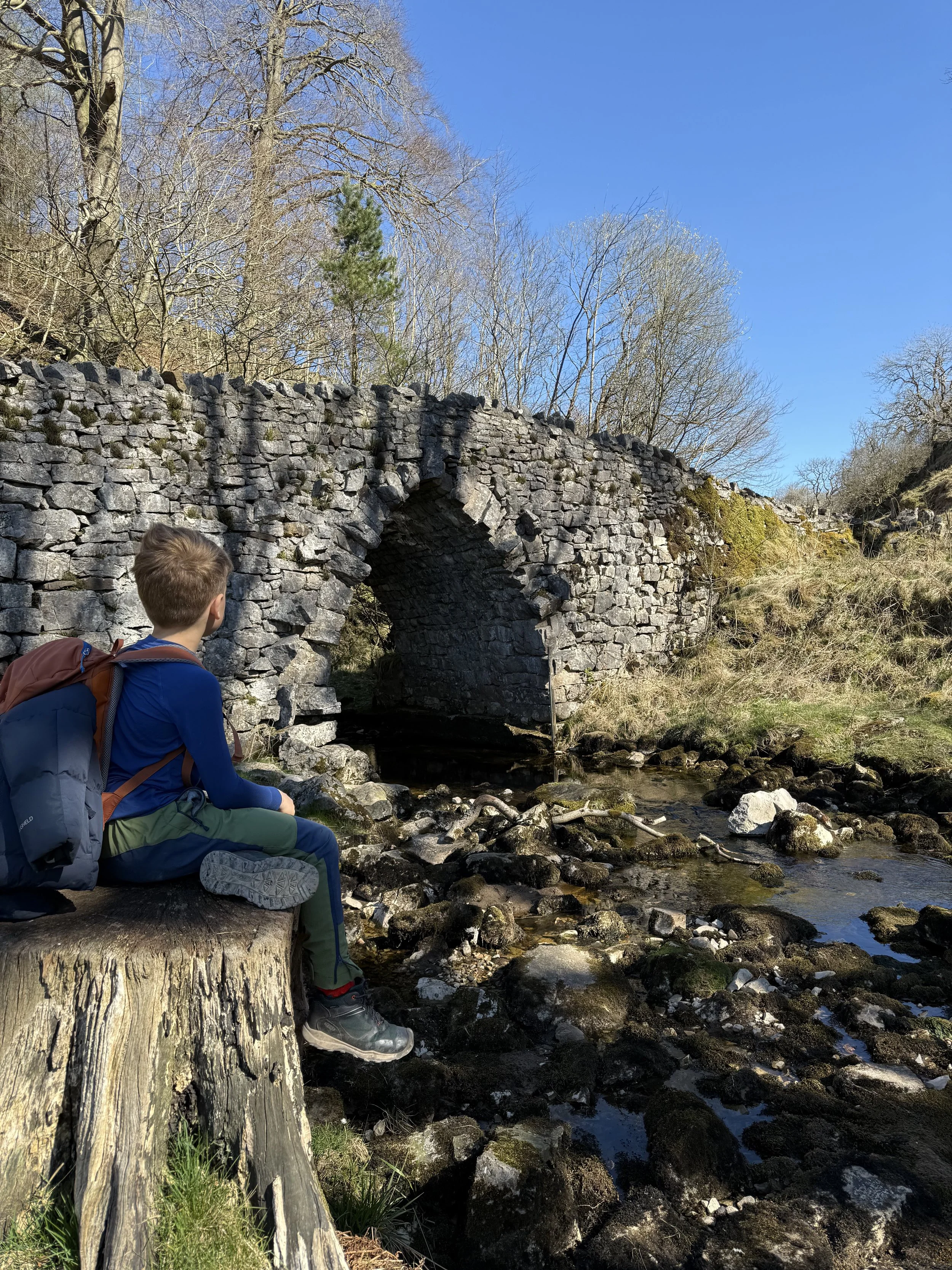 Child sat next to a stone bridge with a river running past under blue spring skies