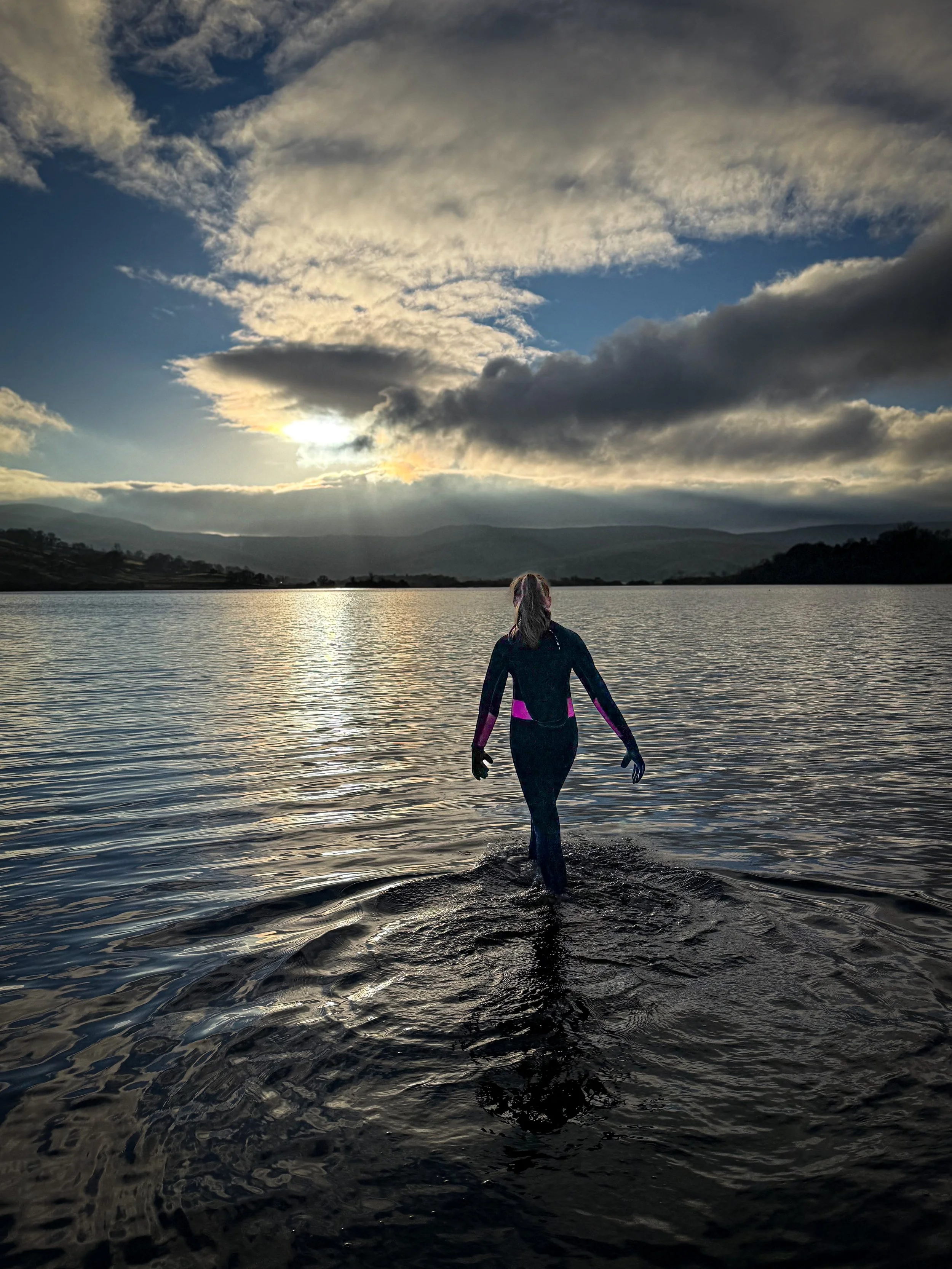 Child in westuit walking into water of lake with low sun behind