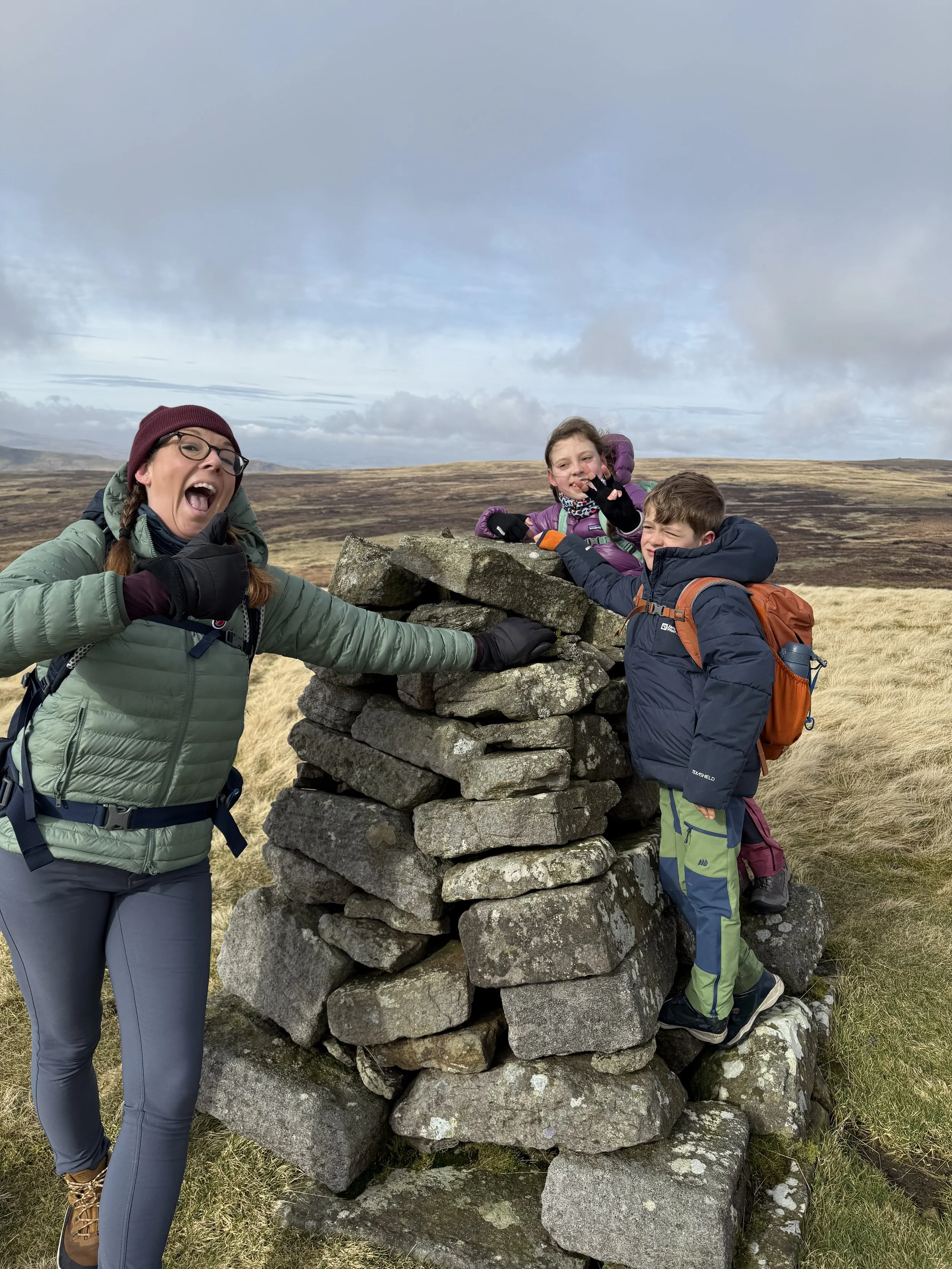 An adult and two children at a stone cairn on top of a mountain with blue skies and grassy fells beyond